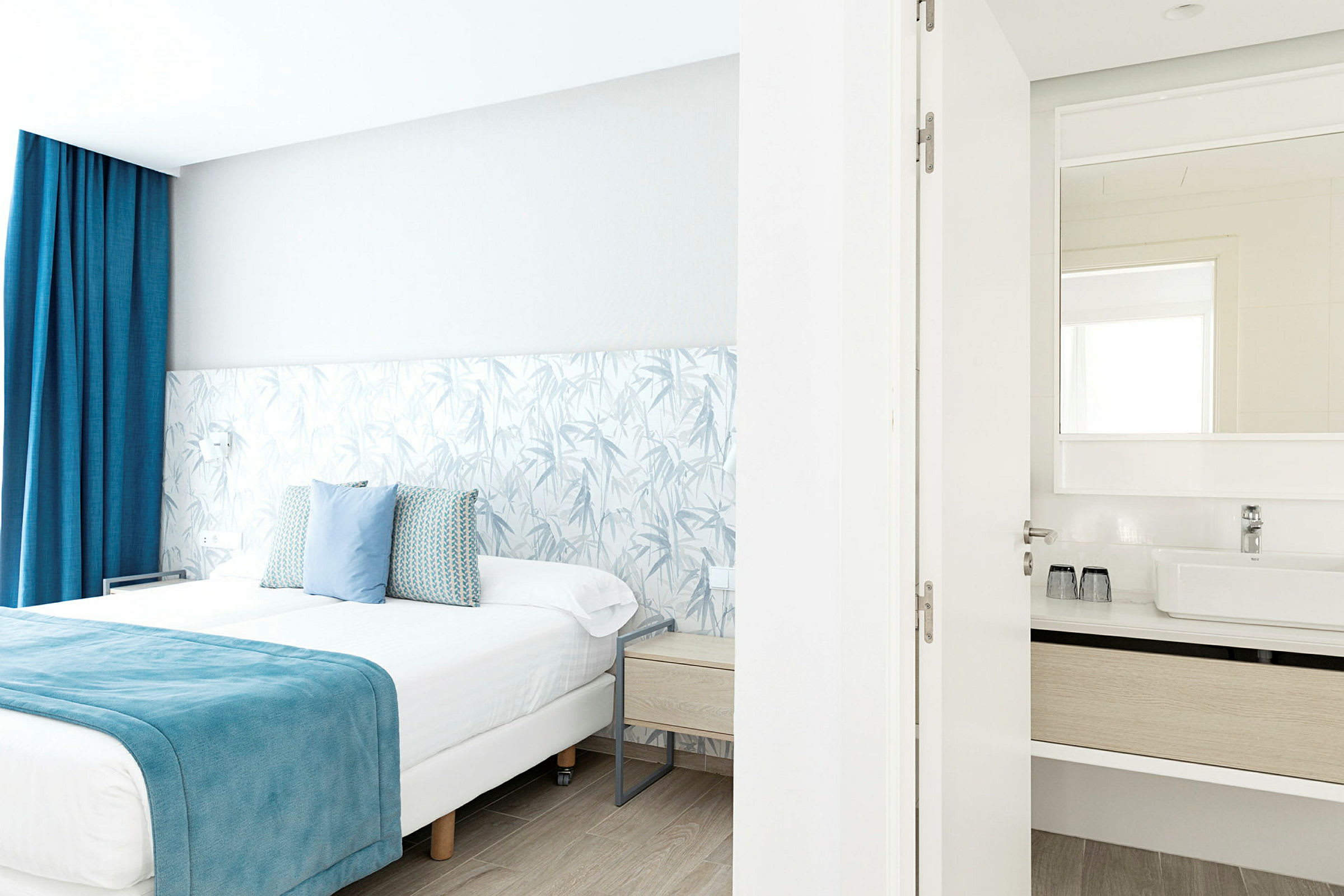 Valoisa hotellihuone, jossa on parivuode sinisin yksityiskohdin, ja liitetty kylpyhuone näkyvällä peilillä. (Tekoälyn luoma teksti)