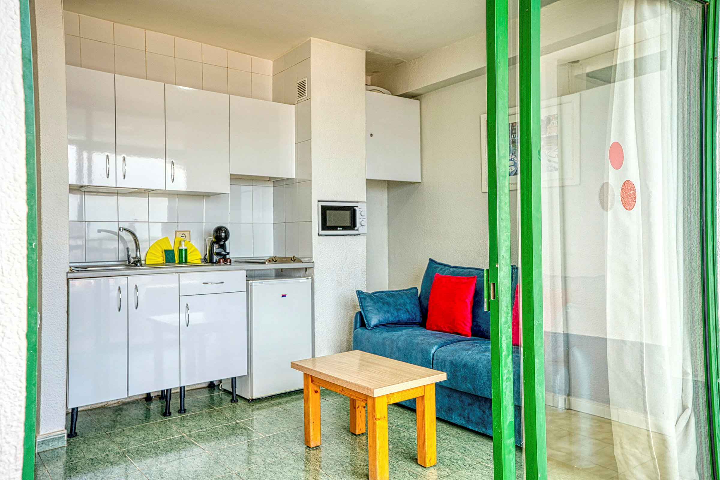 Kompakti huoneisto, jossa pieni keittiö, sininen sohva punaisilla tyynyillä ja yksinkertainen puupöytä, erotettuna lasiovella. (Tekoälyn luoma teksti)
