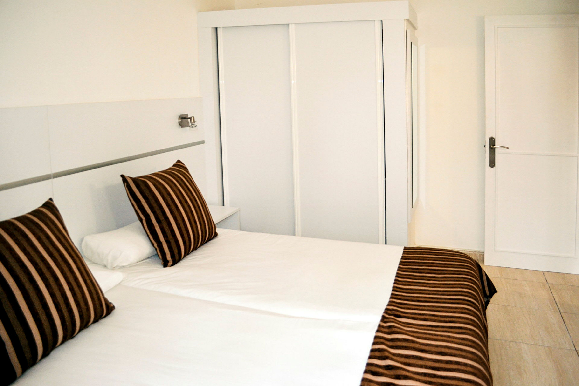 Hotellihuone, jossa valkoinen sänky ja ruskealla raidallisella kuviolla varustetut tyynyt valkoisen vaatekaapin edessä. (Tekoälyn luoma teksti)
