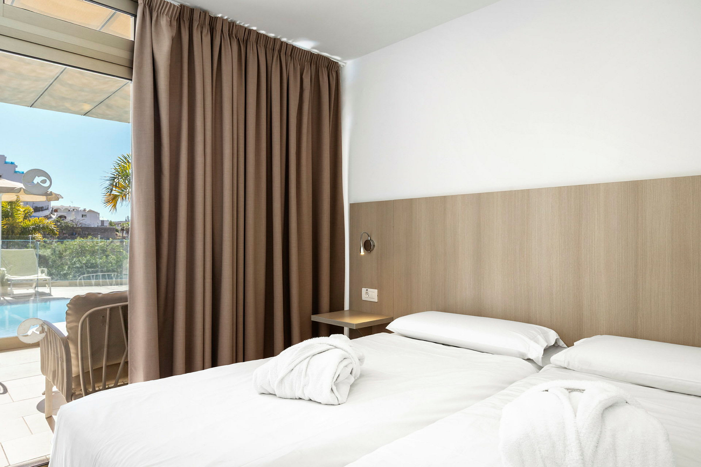Hotellihuone, jossa on kaksi yhden hengen vuodetta ja valkoiset lakanat. Huoneessa on ruskeat verhot ja lasiovi, joka johtaa terassille, jolta on näkymä uima-altaalle. (Tekoälyn luoma teksti)