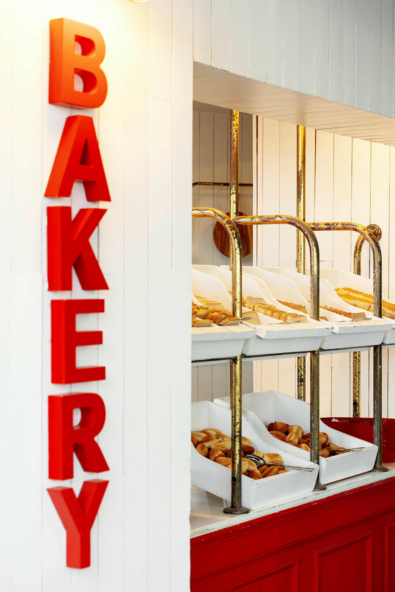 Leipomoalue, jossa on punaisia kirjaimia, jotka muodostavat sanan 'BAKERY'. Hyllyissä on erilaisia leipiä ja leivonnaisia valkoisissa laatikoissa. (Tekoälyn luoma teksti)