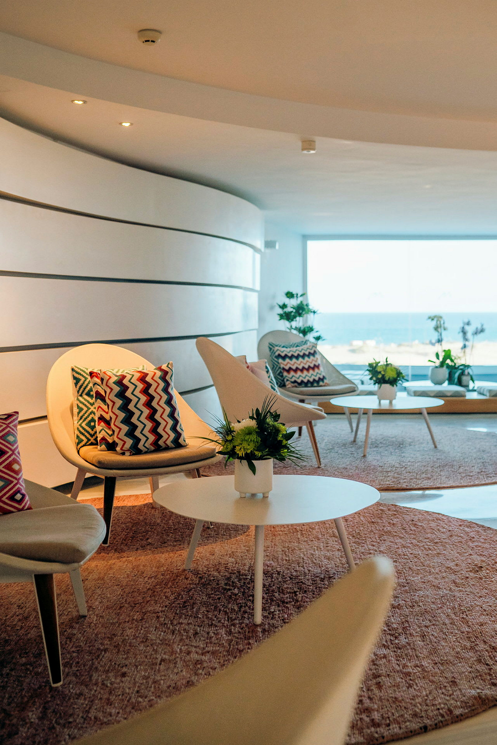 Lounge-tila, jossa on moderneja tuoleja ja sohvapöytiä, koristeltuina värikkäillä tyynyillä ja kukilla, merinäköalalla suuren ikkunan läpi. (Tekoälyn luoma teksti)