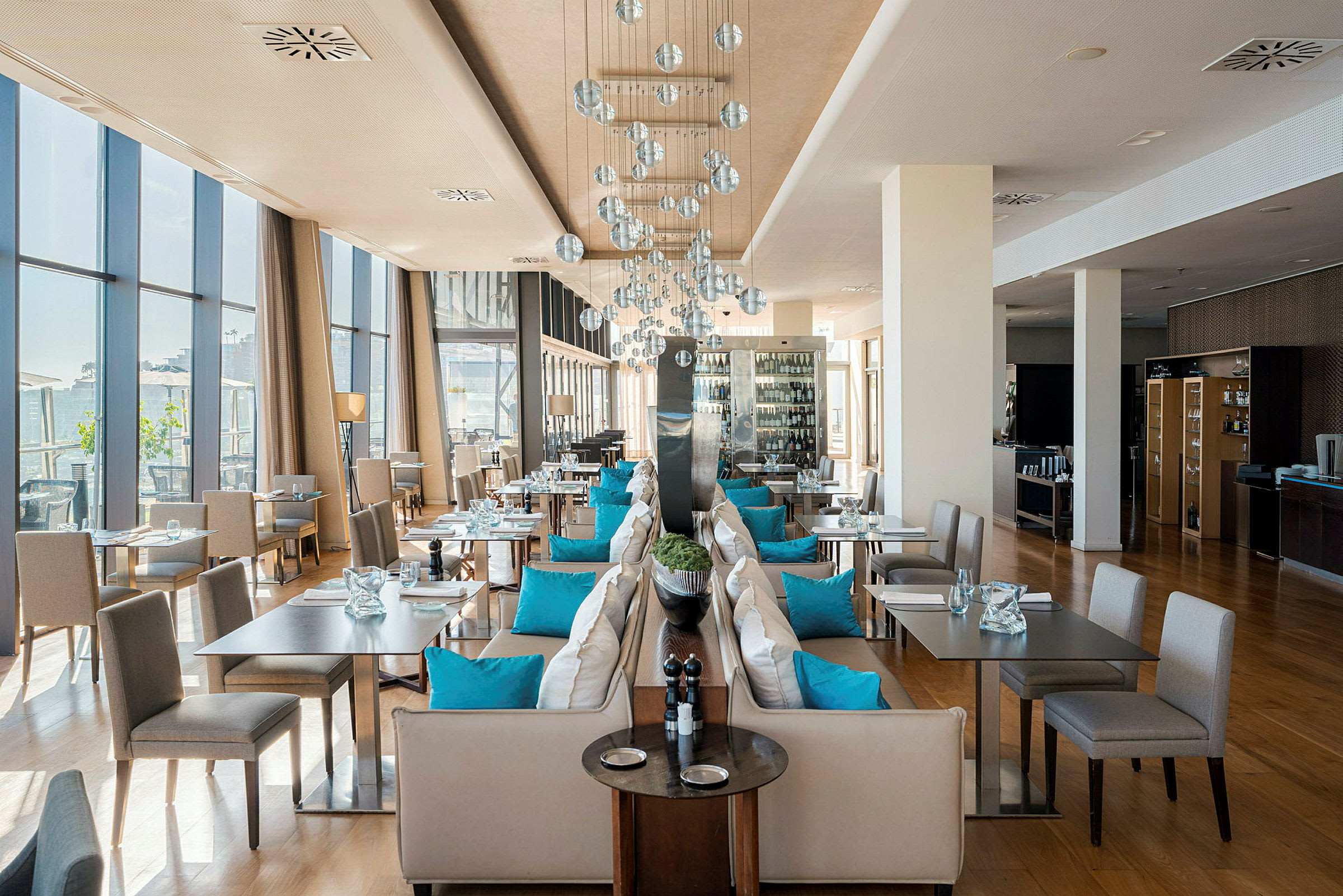 Tyylikäs ravintola, jossa on suuret ikkunat, sinisillä tyynyillä koristeltuja penkkejä ja modernit valaisimet. (Tekoälyn luoma teksti)