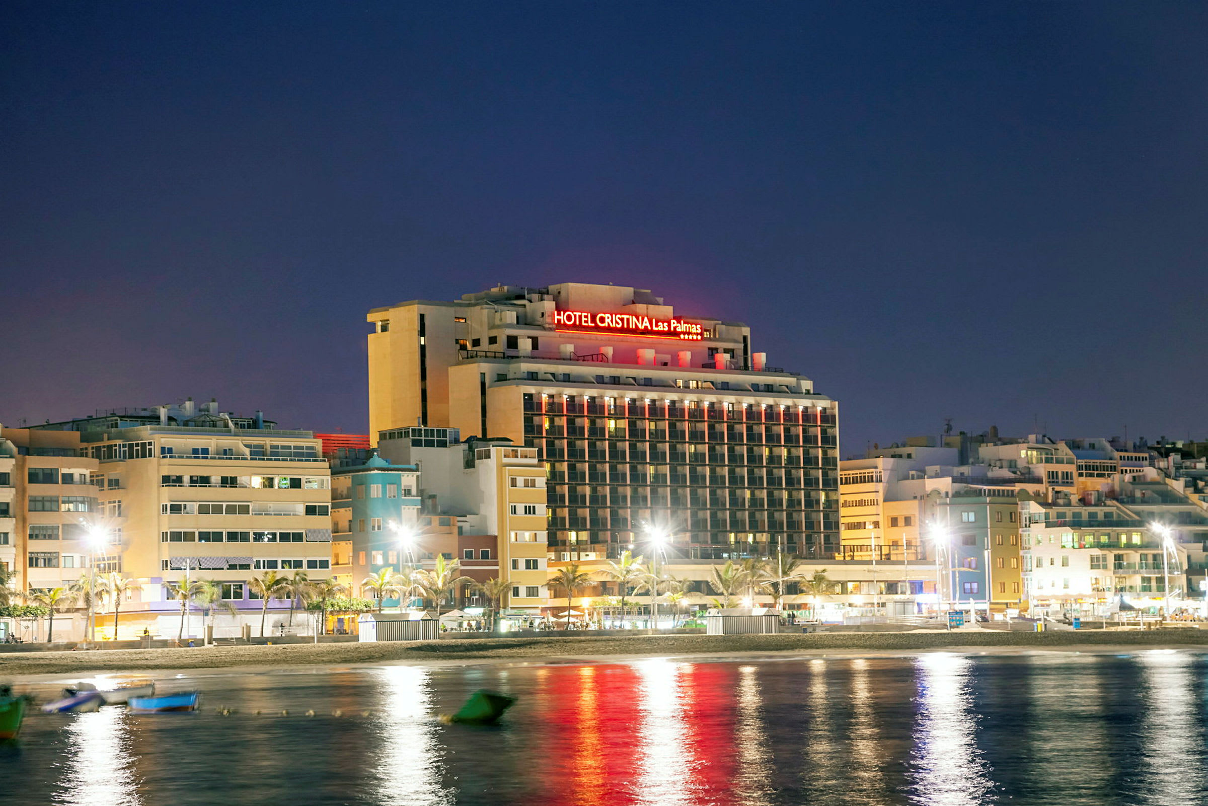 Kuva hotellirakennuksesta neonvaloilla yöllisessä ympäristössä, sijaitsee veden lähellä. (Tekoälyn luoma teksti)
