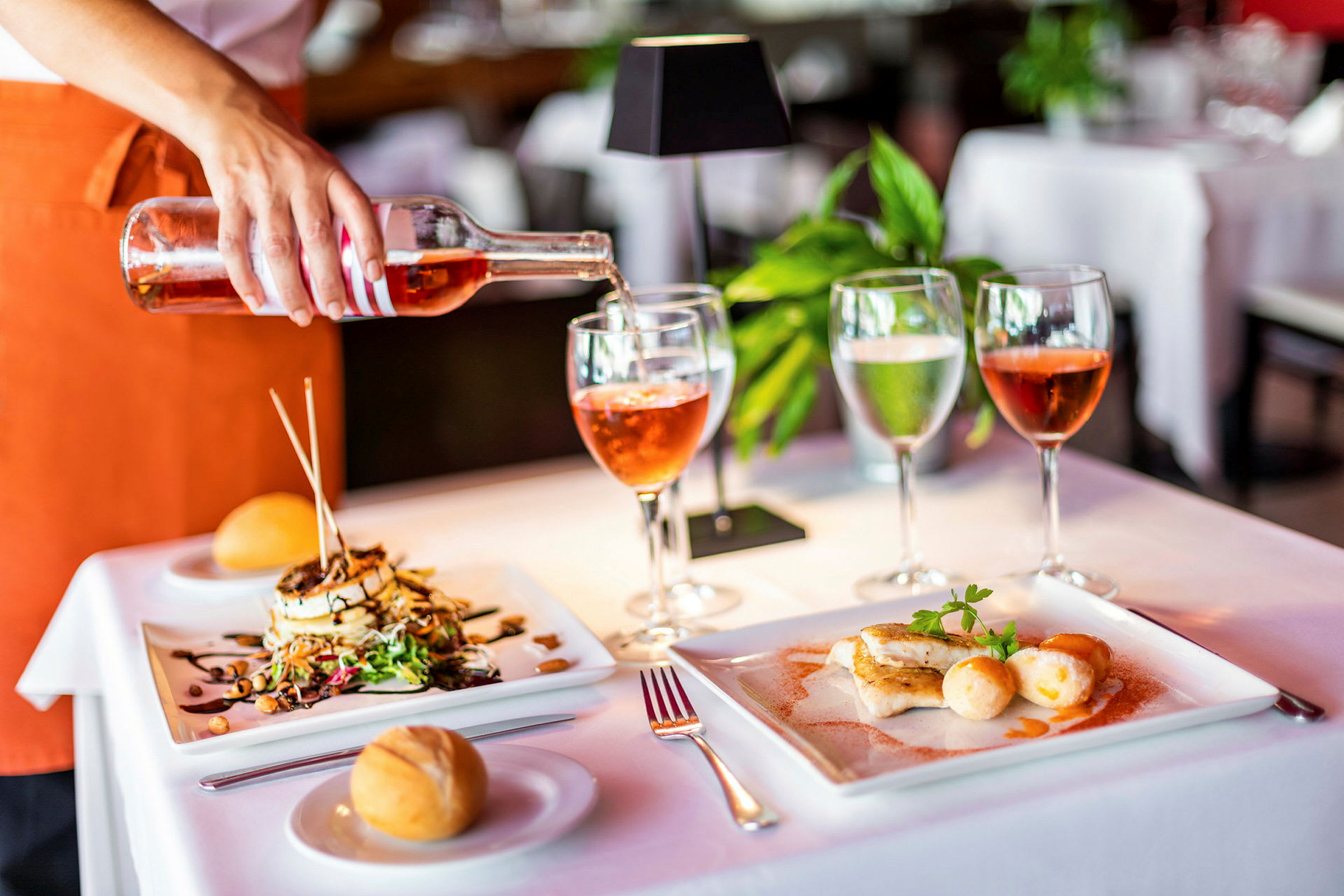 Pöytä katettu lautasilta ruoalla, lasit ja leipä, sekä käsi kaatamassa rosé-viiniä. (Tekoälyn luoma teksti)