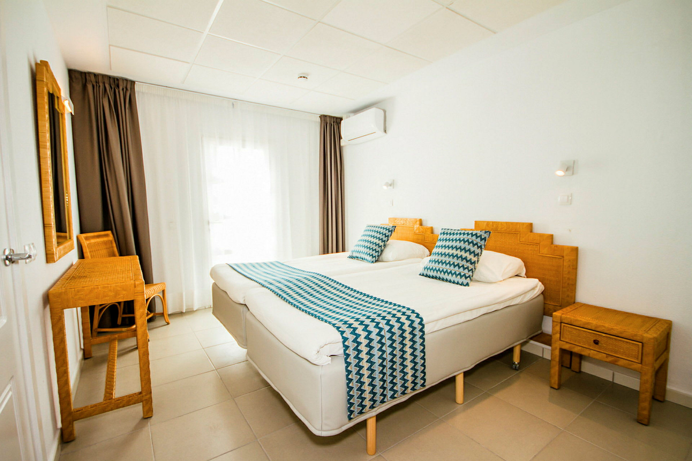 Hotellihuone, jossa kaksi yhden hengen vuodetta, bambukalusteet ja valkoiset seinät. (Tekoälyn luoma teksti)