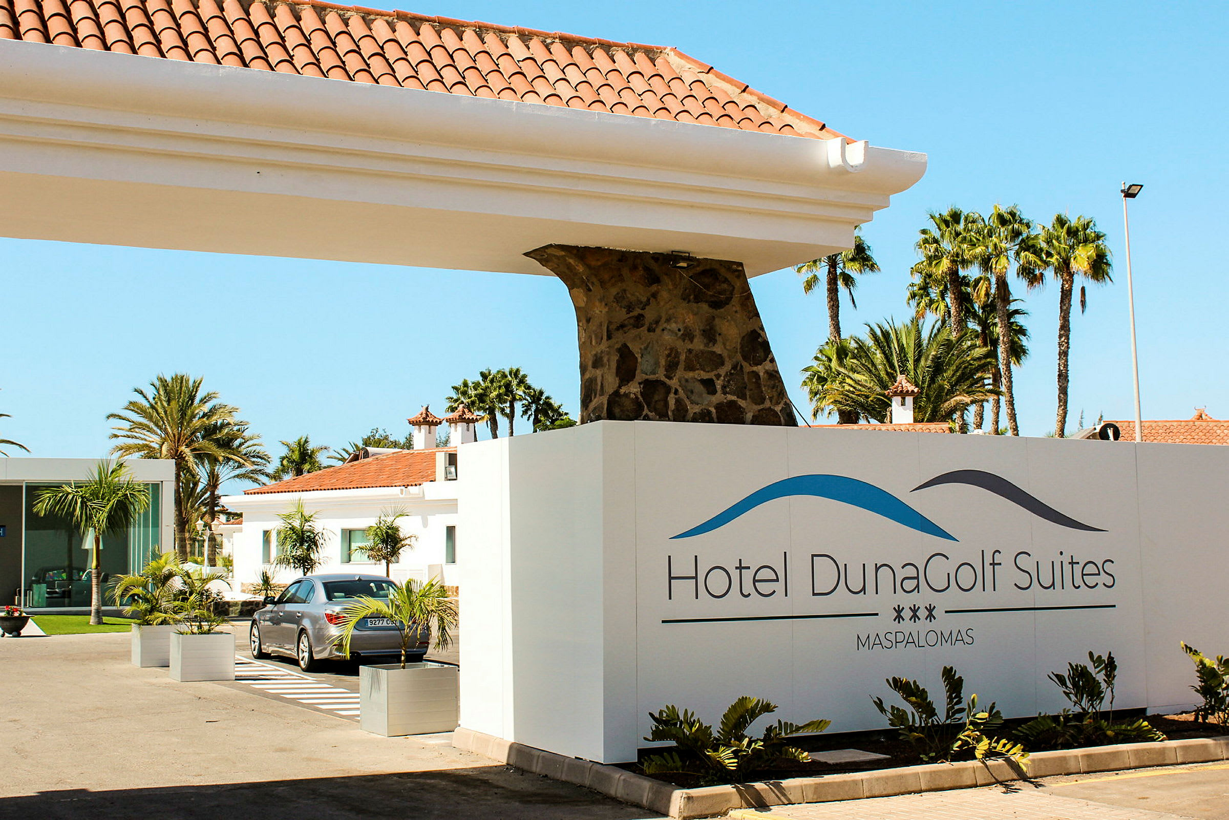 Hotel DunaGolf Suitesin sisäänkäynti, jossa näkyy kyltti ja taustalla rakennuksia. (Tekoälyn luoma teksti)