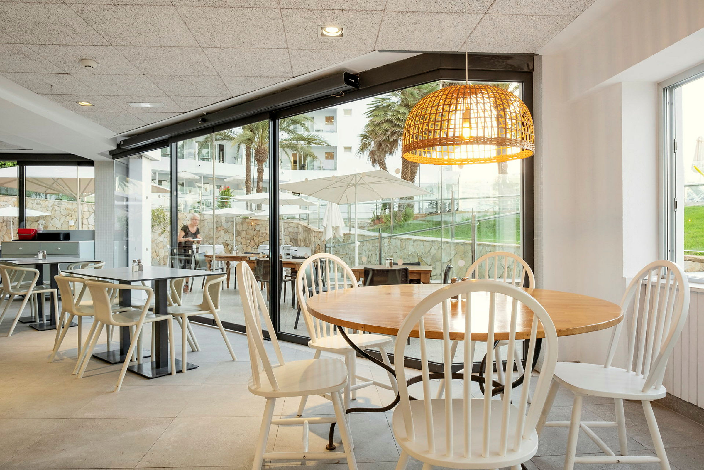 Ravintola, jossa pöytiä ja tuoleja. Suuret lasi-ikkunat näyttävät näkymän, jossa on aurinkovarjoja, vehreitä alueita ja palmuja. (Tekoälyn luoma teksti)