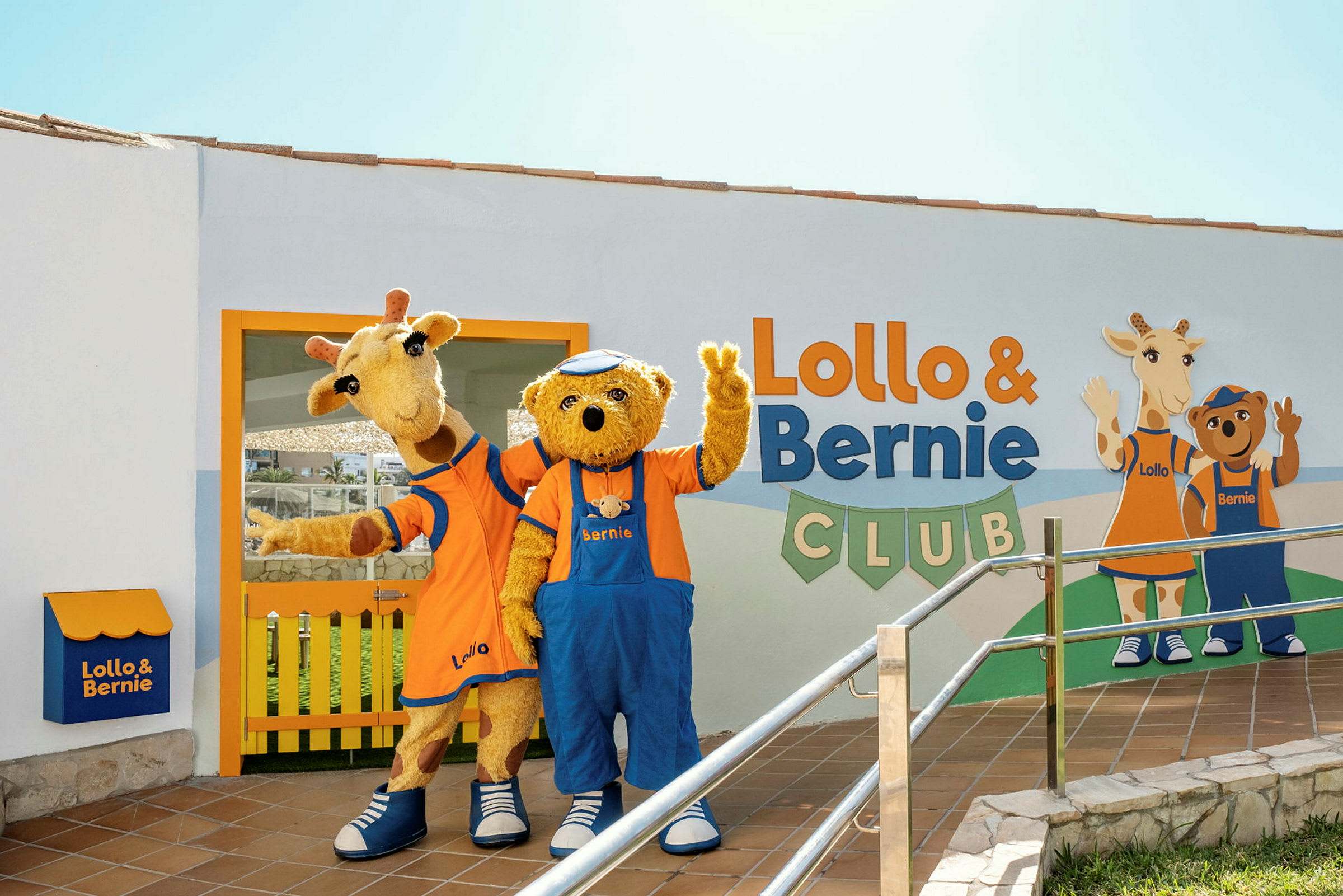 Kaksi maskottia, kirahvi ja karhu, seisovat Lollo & Bernie Clubin rakennuksen edessä ja vilkuttavat. (Tekoälyn luoma teksti)