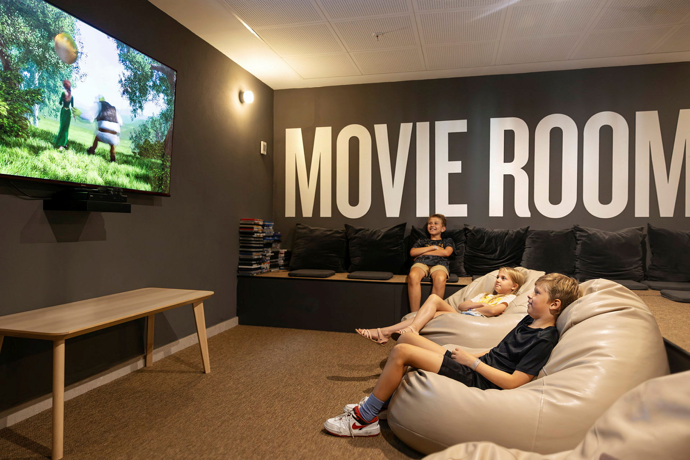 Huone, jossa on pehmeät istuimet ja suuri TV, joka näyttää animaatioelokuvan. Seinissä on teksti "MOVIE ROOM". (Tekoälyn luoma teksti)
