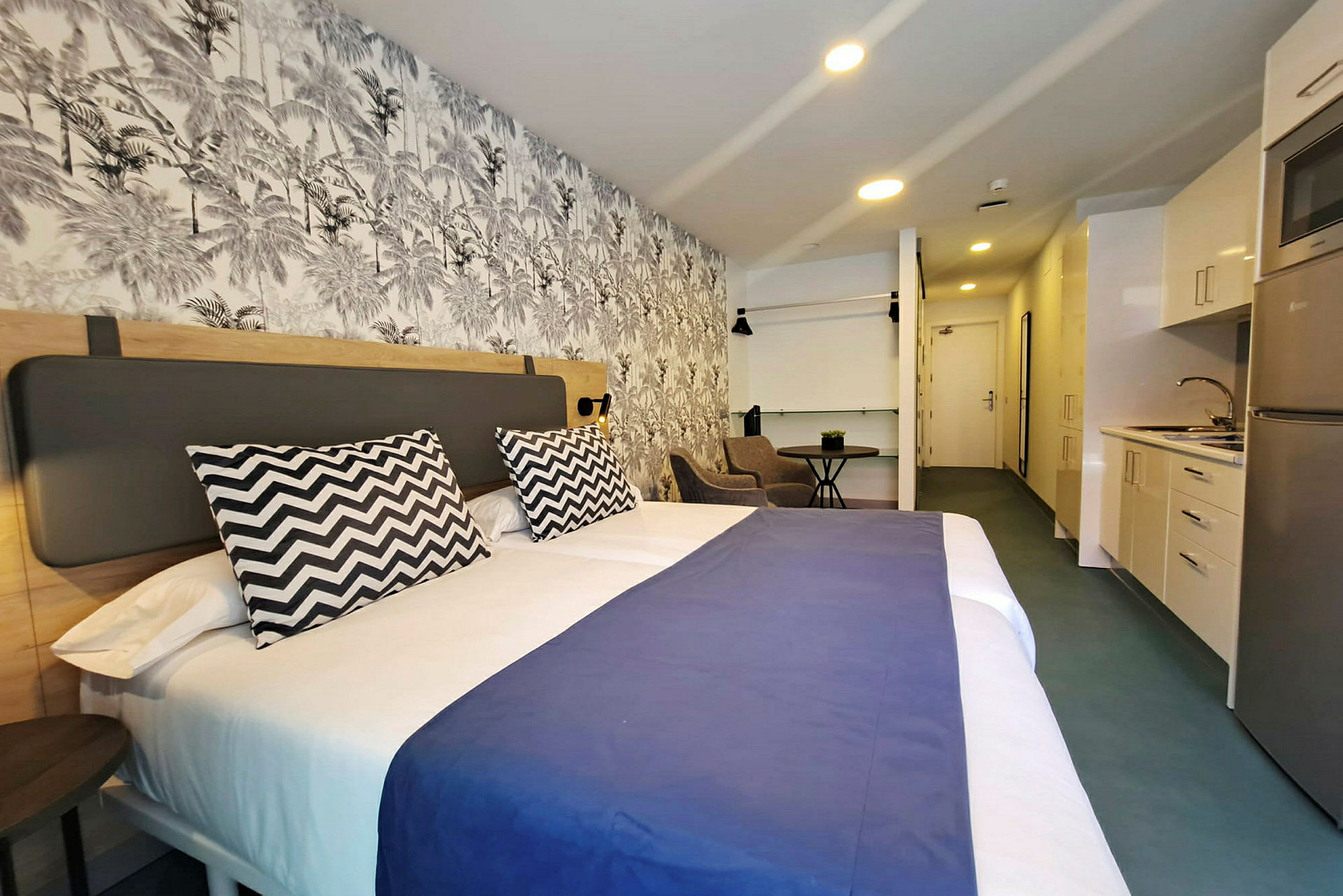 Hotellihuone, jossa suuri sänky, koristeellinen seinä ja pieni keittiö. (Tekoälyn luoma teksti)