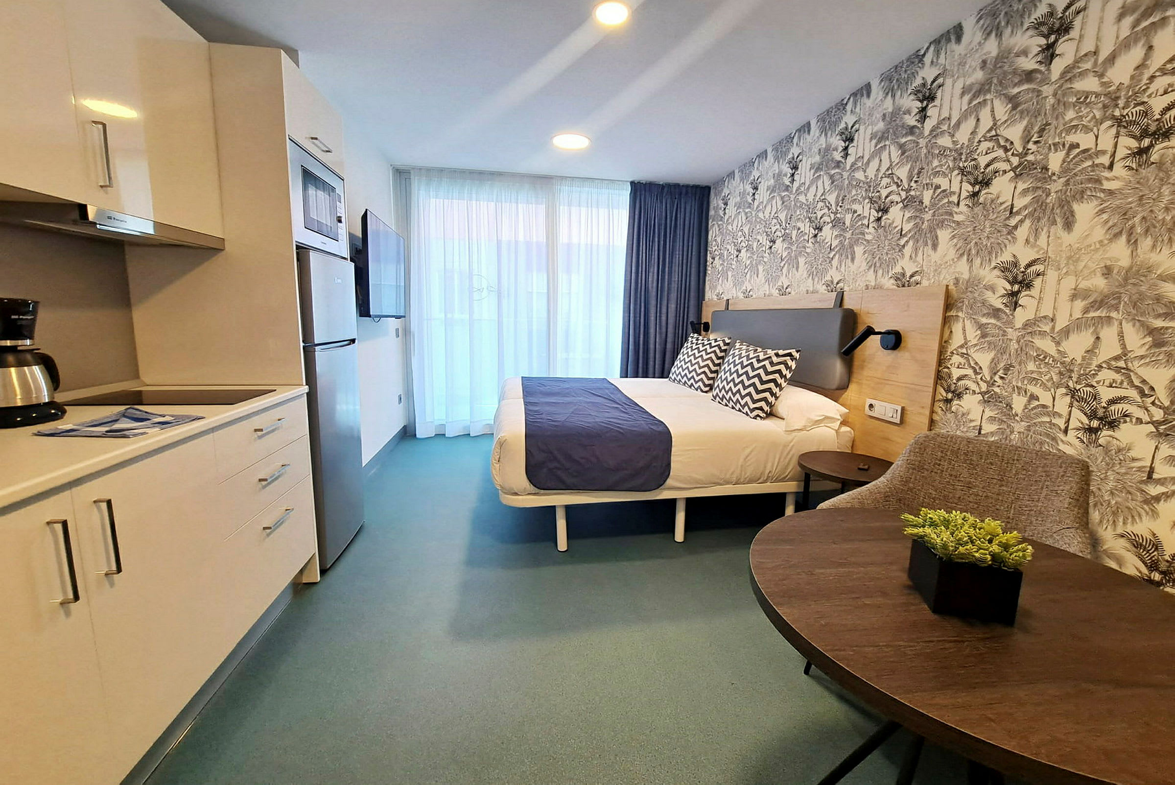 Hotellihuone, jossa on parivuode, keittonurkkaus ja istumisalue. Seinät ovat kuviolliset tapetit. (Tekoälyn luoma teksti)