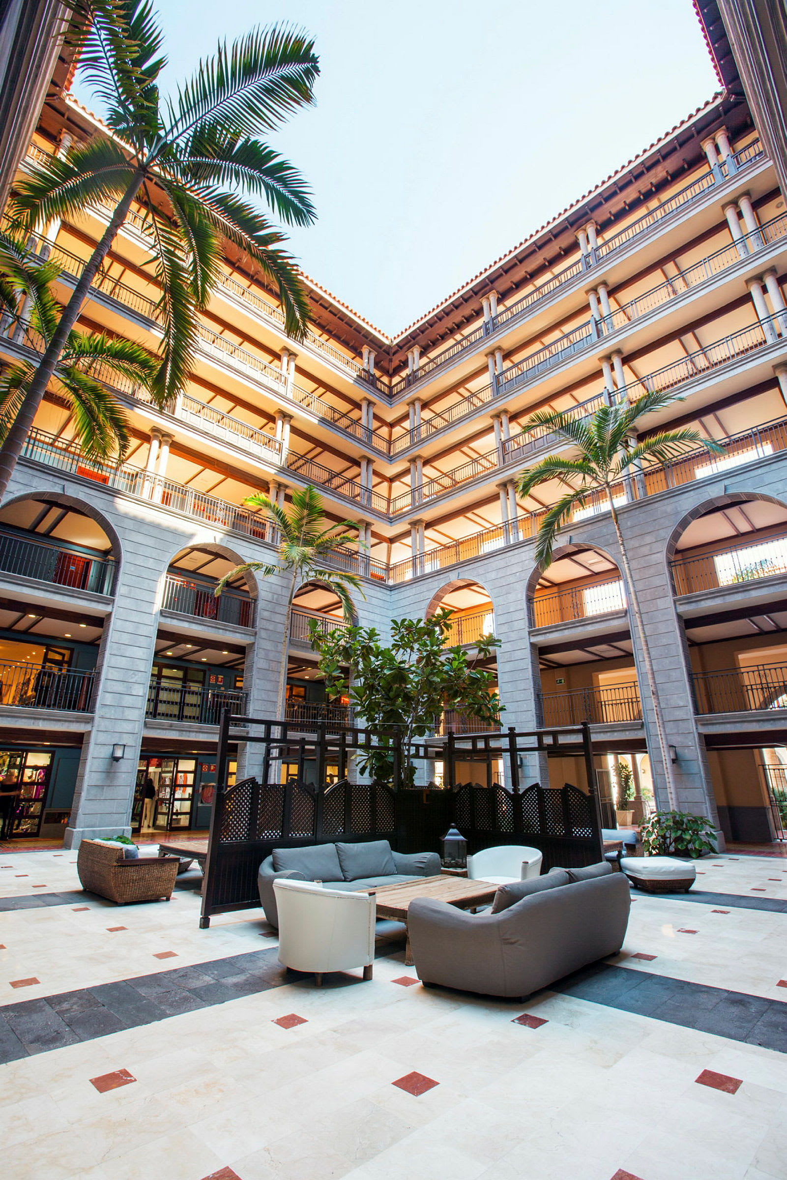 Valoisa atrium, jossa on istumapaikat keskellä, ympäröitynä useilla parvekekerroksilla ja korkeilla palmuilla. (Tekoälyn luoma teksti)