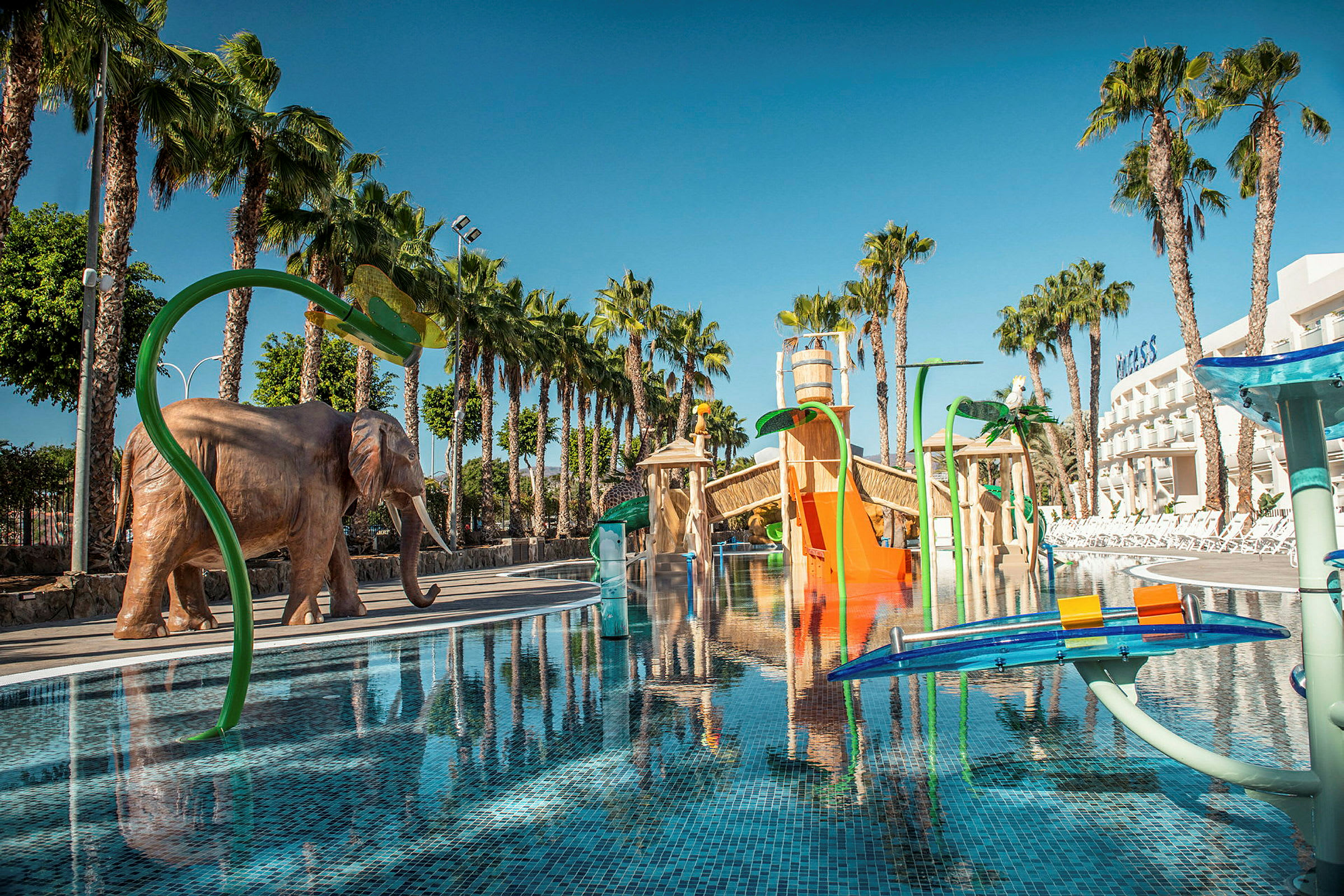 Ulkoilma vesileikkipuisto, jossa on uima-allas, värikkäitä vesiliukumäkiä ja suuri norsupatsas, palmujen ympäröimänä. (Tekoälyn luoma teksti)