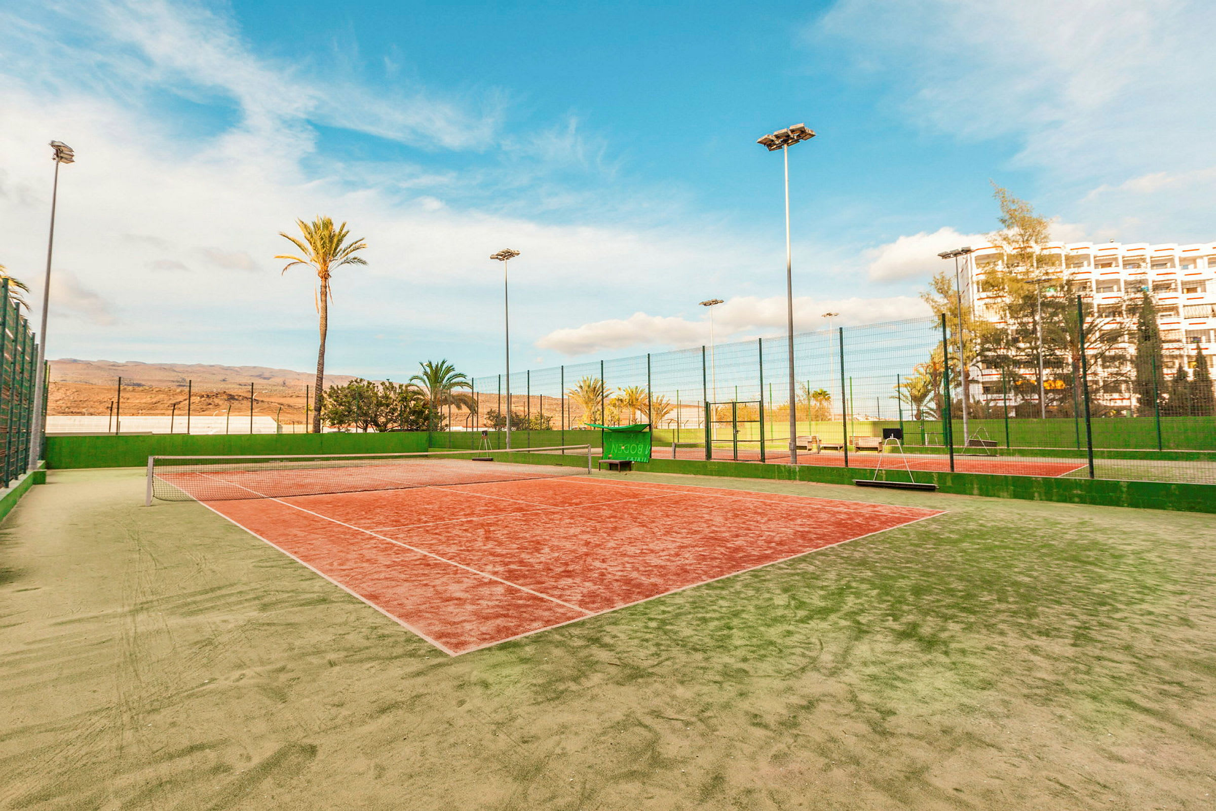 Tenniskenttä vihreällä ja punaisella pinnalla, jota ympäröivät korkeat verkot ja palmut. (Tekoälyn luoma teksti)