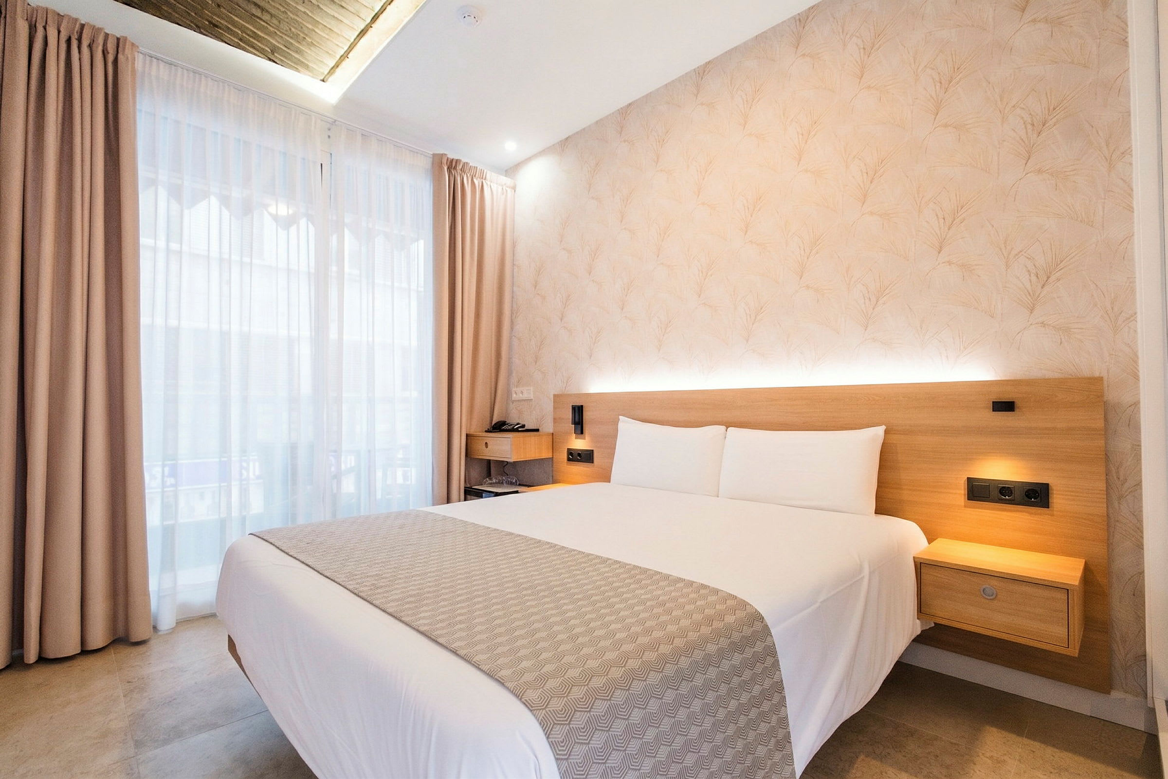 Hotellihuoneessa on parivuode, vaalea päiväpeite ja tyynyjä. Vaalea seinä, jossa on puupaneeli, ja pieni yöpöytä ovat näkyvissä. Ikkunat, joissa on vaaleat verhot, päästävät sisään päivänvaloa. (Tekoälyn luoma teksti)