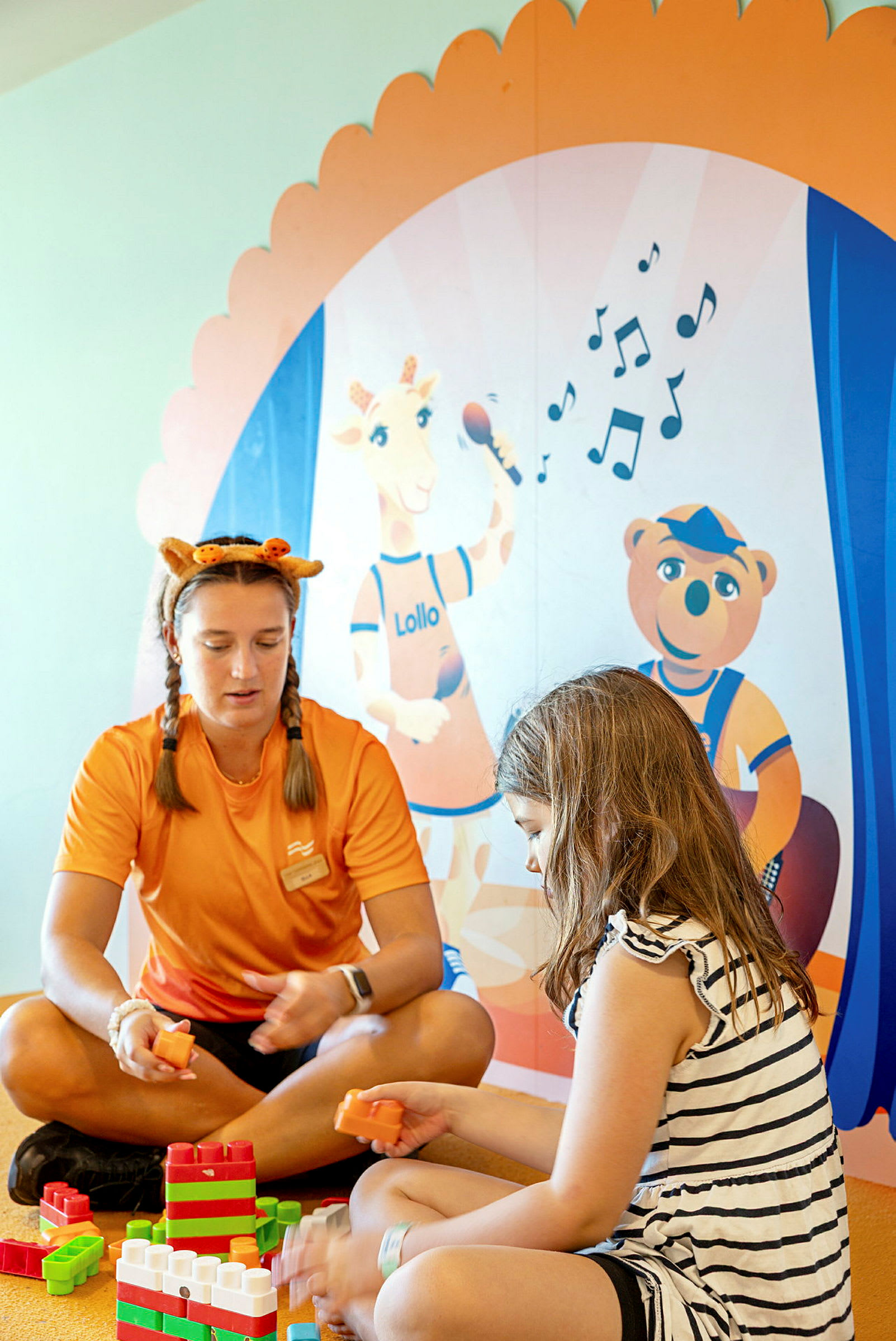 Huone, jossa on lasten leluja, jossa kaksi henkilöä leikkii rakennuspalikoilla värikkään seinän edessä, jolla on lasten hahmokuvia. (Tekoälyn luoma teksti)
