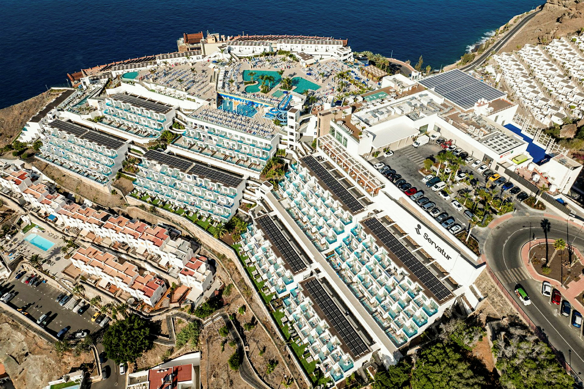 Ilmakuva suuresta hotellikompleksista meren rannalla, jossa on allasalue ja ympäröiviä asuinrakennuksia. (Tekoälyn luoma teksti)