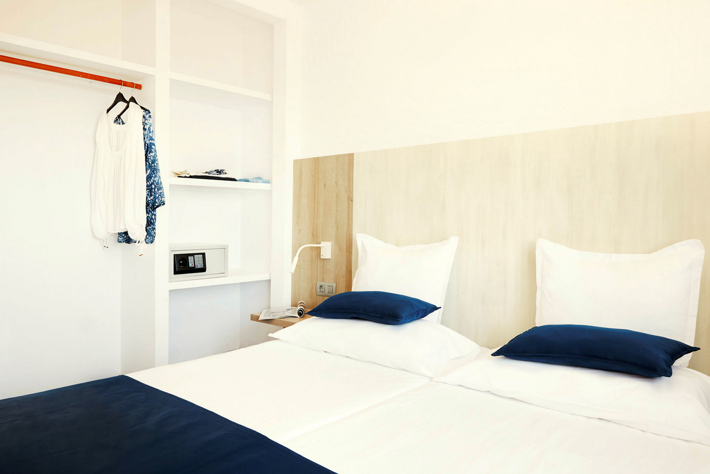 Hotellihuone, jossa on kaksi sänkyä valkoisilla ja sinisillä vuodevaatteilla, vaatekaappi vaatteineen ja tallelokero. (Tekoälyn luoma teksti)