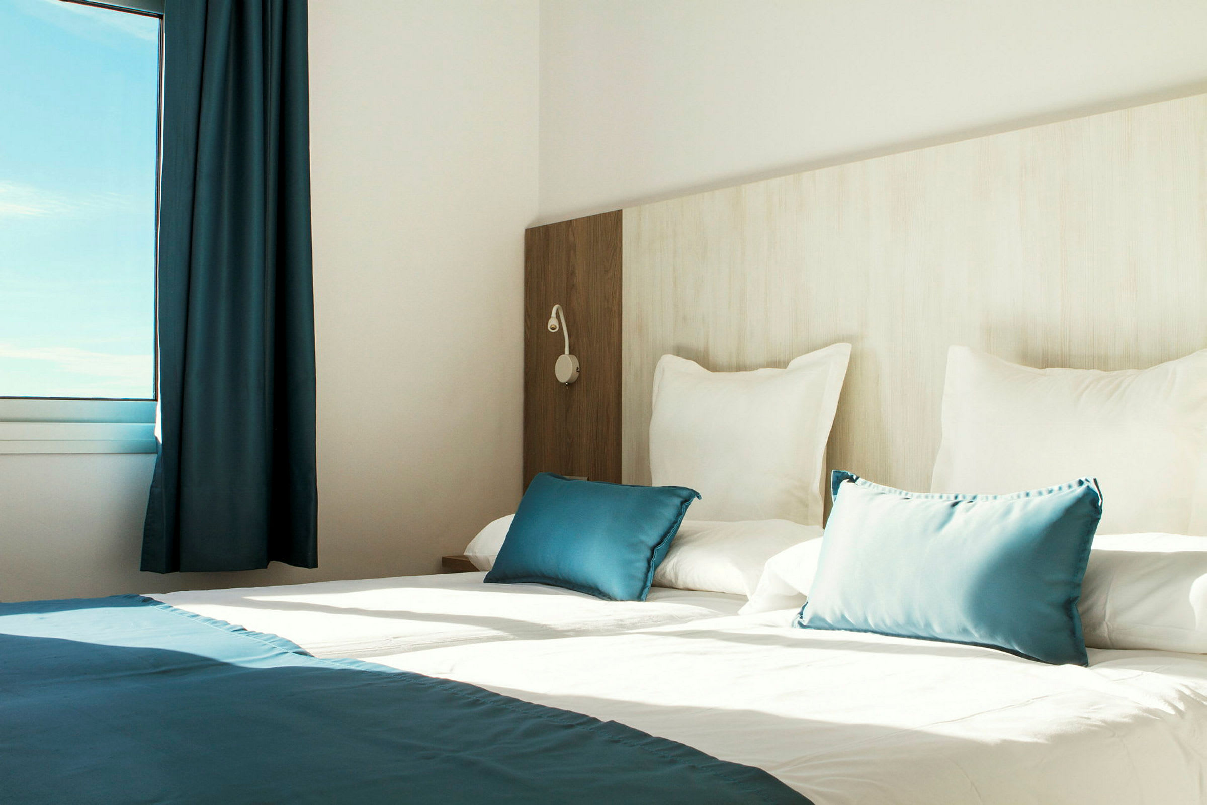 Valoisan hotellihuoneen sisustus, jossa on pedattu sänky, valkoisia ja sinisiä tyynyjä sekä ikkuna, josta tulee luonnonvaloa. (Tekoälyn luoma teksti)