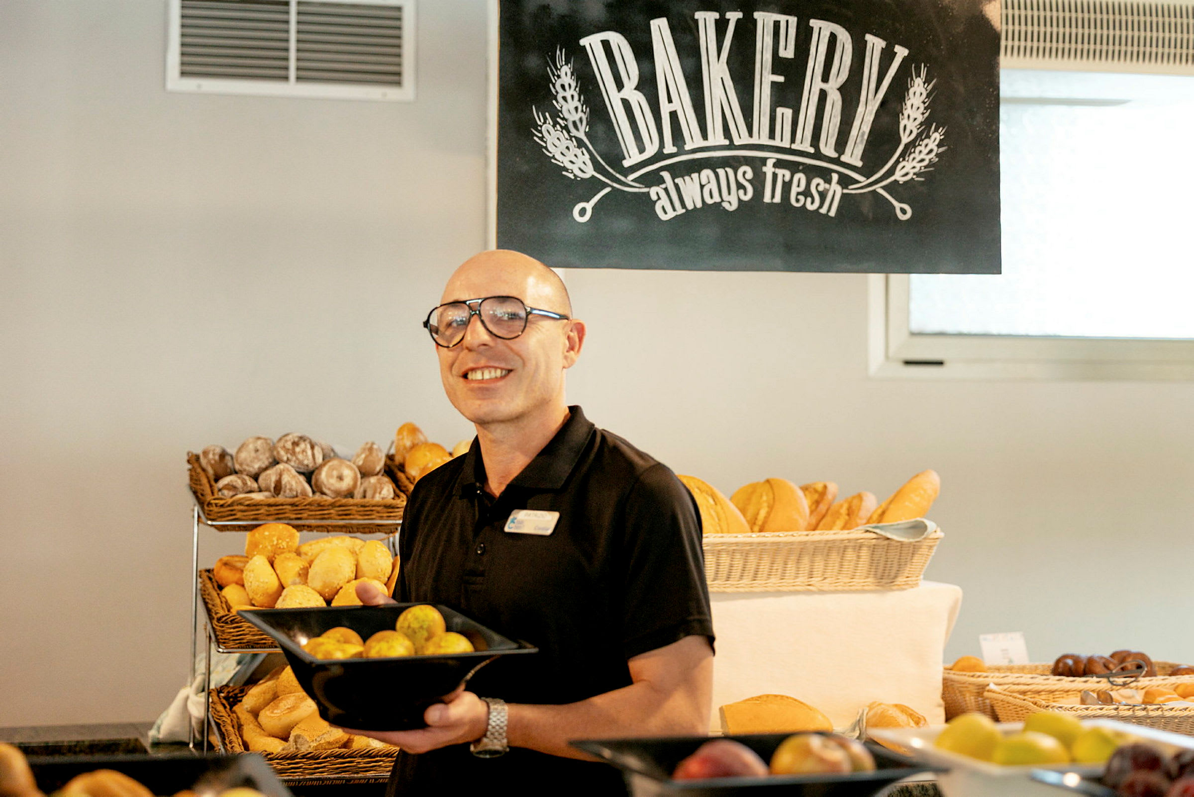 Henkilö pitää mustaa kulhoa, jossa on leipää, leipomossa. Taustalla näkyy muita leivonnaisia ja kyltti 'Bakery always fresh' -tekstillä. (Tekoälyn luoma teksti)