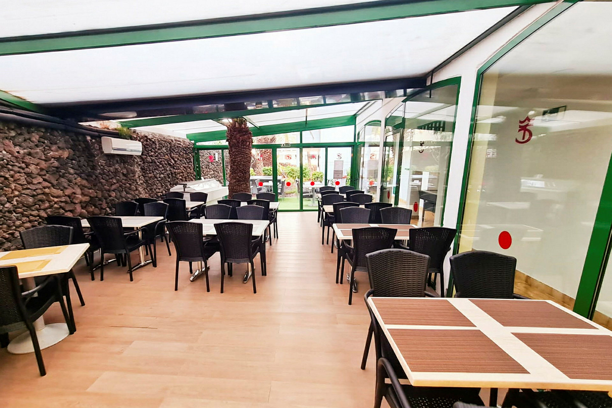 Huone, jossa on useita pöytiä ja tuoleja, lasiseinät ja kivimuuri. (Tekoälyn luoma teksti)