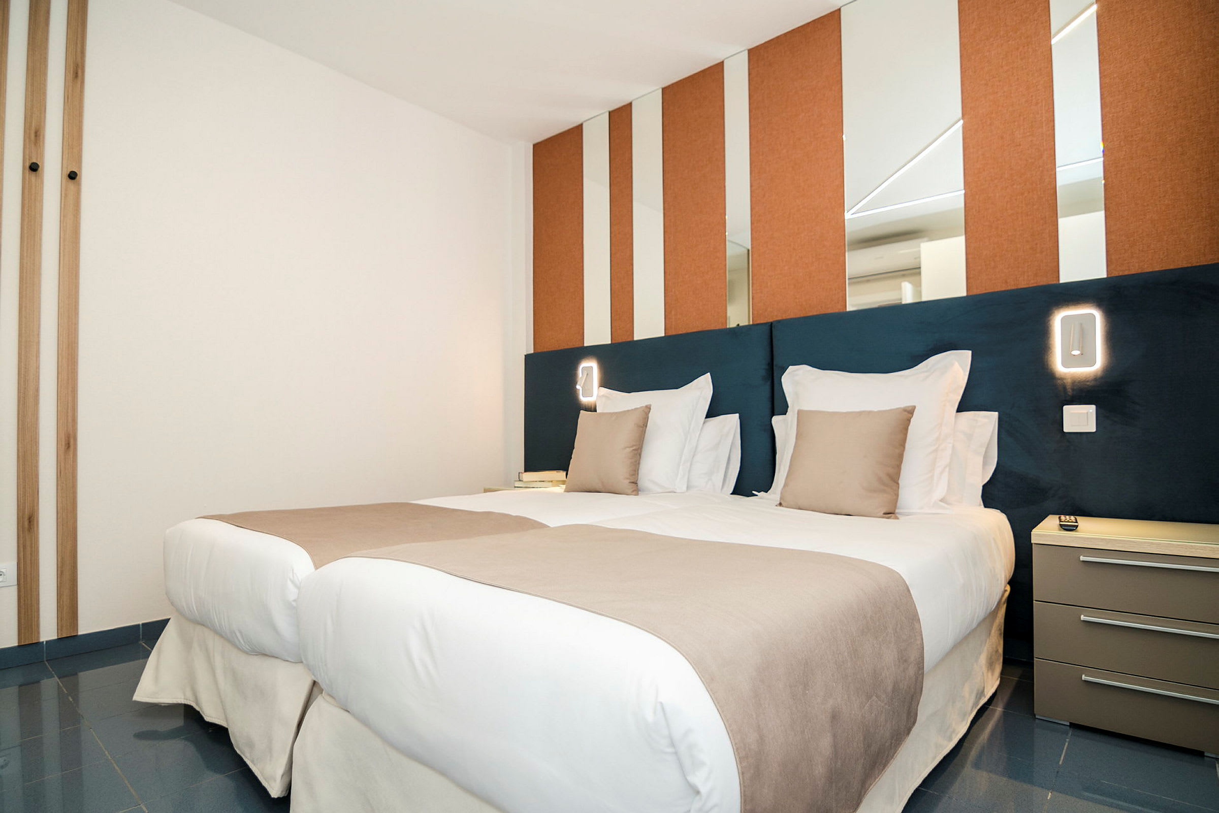 Moderni hotellihuone, jossa kaksi vierekkäistä yhden hengen vuodetta, pedattuna valkoisilla ja beigenvärisillä vuodevaatteilla. Seinissä on peilejä ja oransseja paneeleita, vuoteiden vieressä yöpöytä. (Tekoälyn luoma teksti)