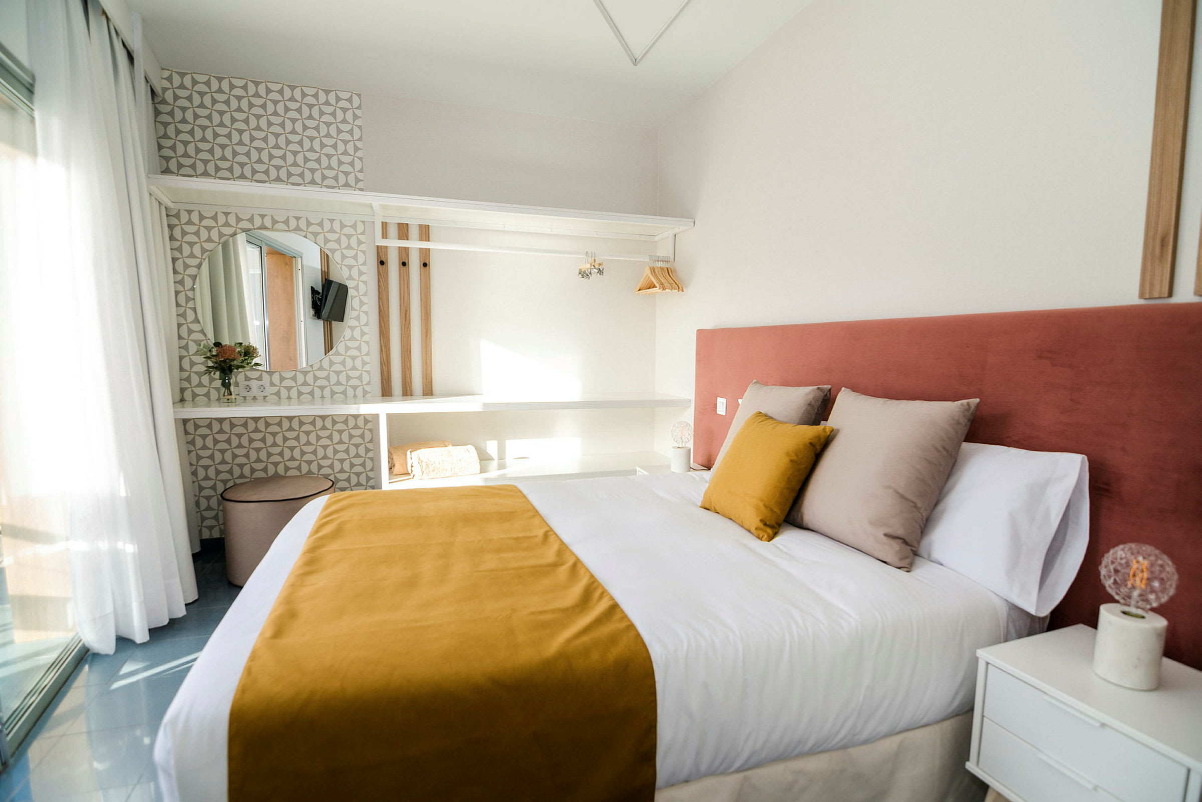 Hotellihuone, jossa on parivuode, keltainen peitto ja vaaleanpunainen sängynpääty, sekä moderni sisustus hyllyillä, peilillä ja valaistuksella. (Tekoälyn luoma teksti)