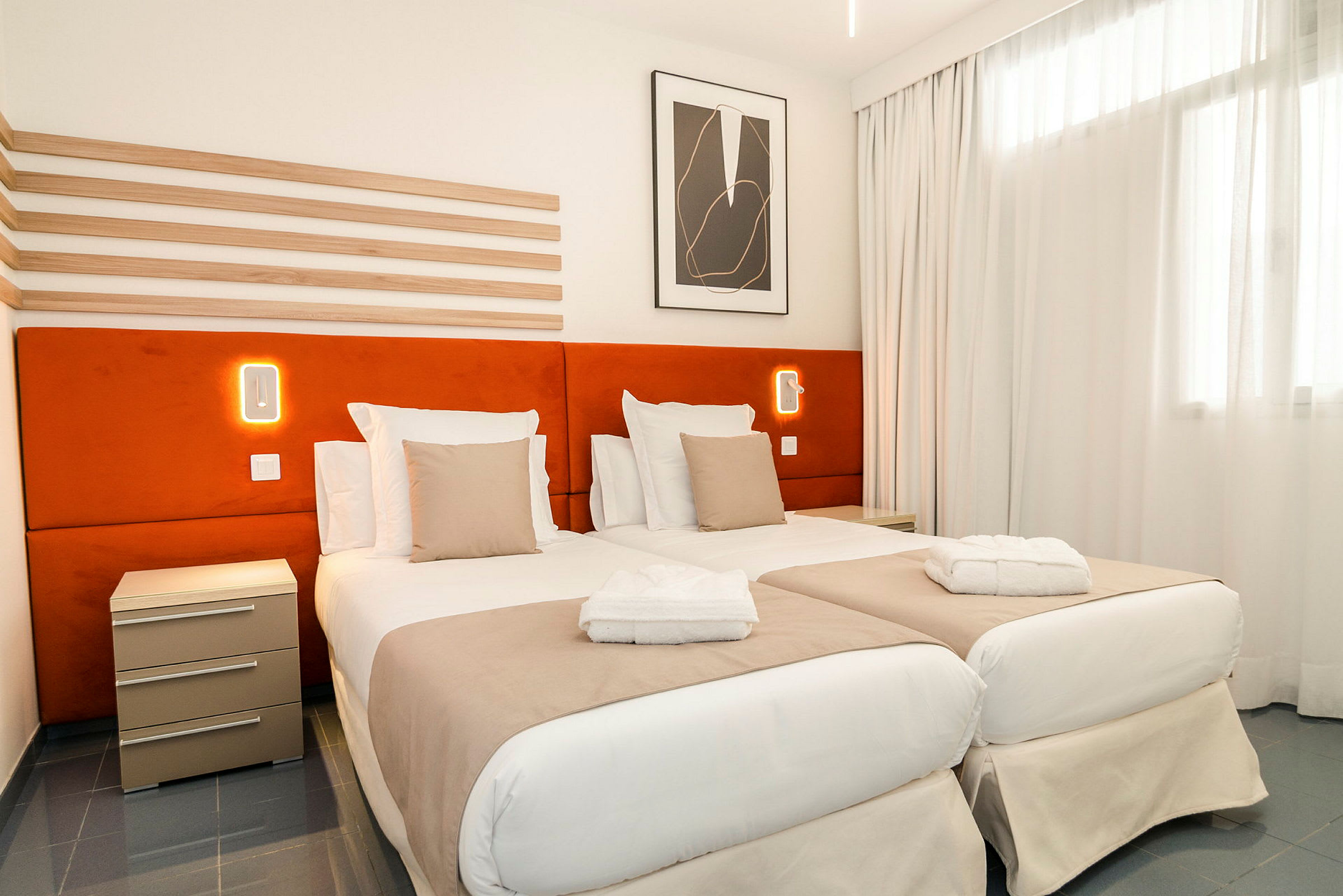 Hotellihuone, jossa on kaksi erillistä sänkyä, oranssit sängynpäädyt ja valkoiset vuodevaatteet. Seinällä näkyy yövalo. (Tekoälyn luoma teksti)