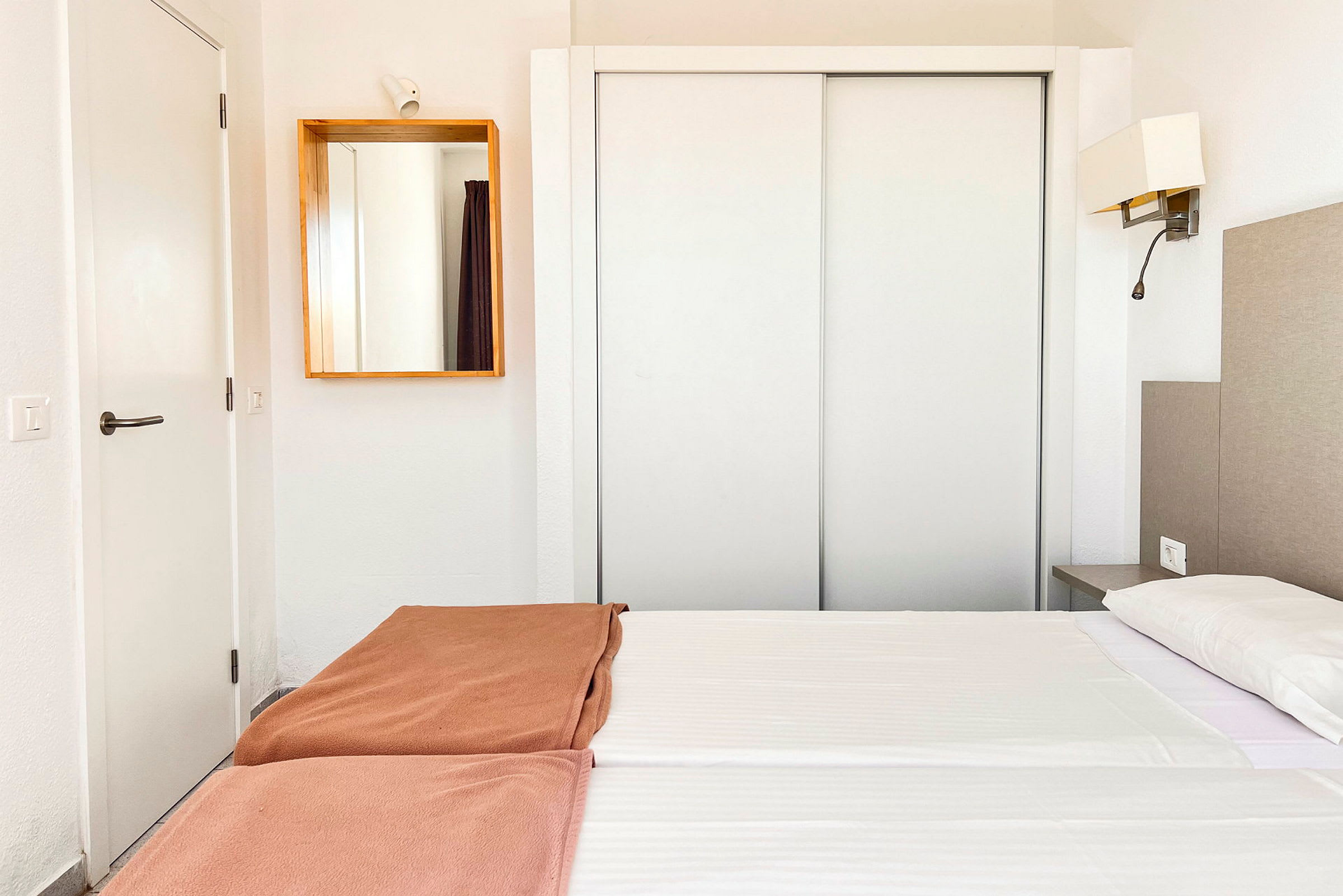 Hotellihuone, jossa valkoiset sisusteet, parivuode valkoisilla lakanoilla ja ruskeilla peitteillä sekä peili seinällä. (Tekoälyn luoma teksti)