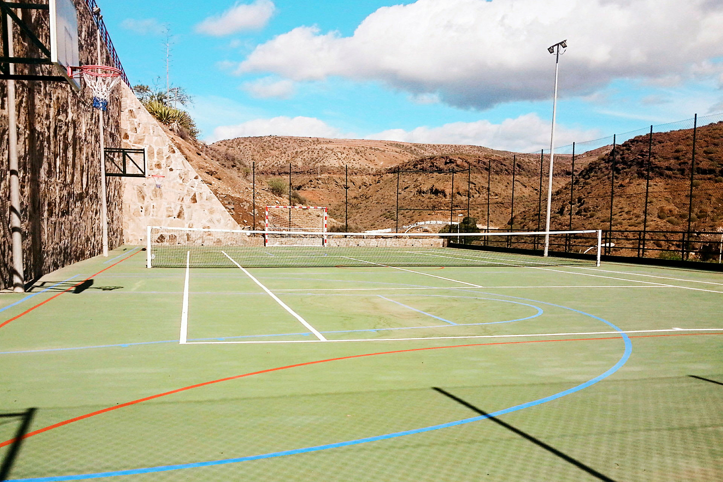 Ulkoilmaurheilukentän kuva, jossa on verkkoja tennistä varten, jalkapallomaali ja koripallokori. (Tekoälyn luoma teksti)