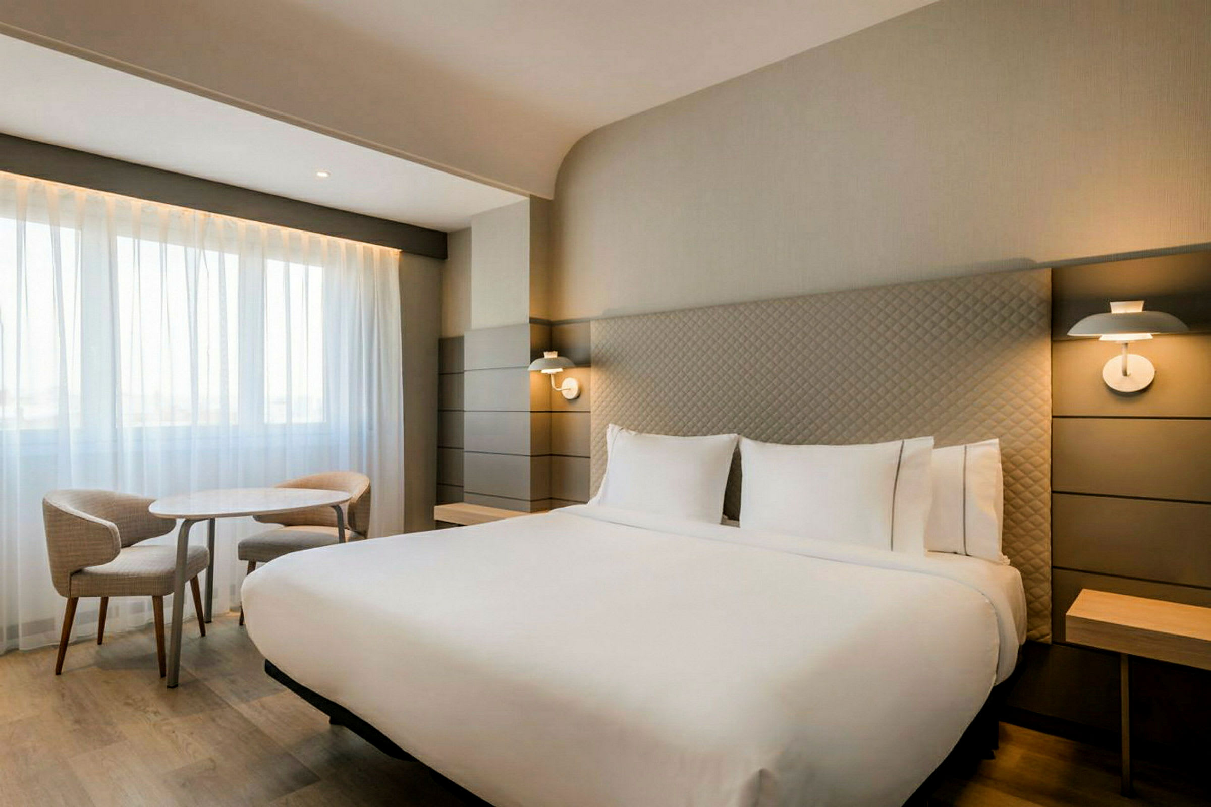 Moderni hotellihuone, jossa on suuri parisänky, kaksi lukulamppua, pöytä ja kaksi tuolia ikkunan ääressä vaaleilla verhoilla. (Tekoälyn luoma teksti)