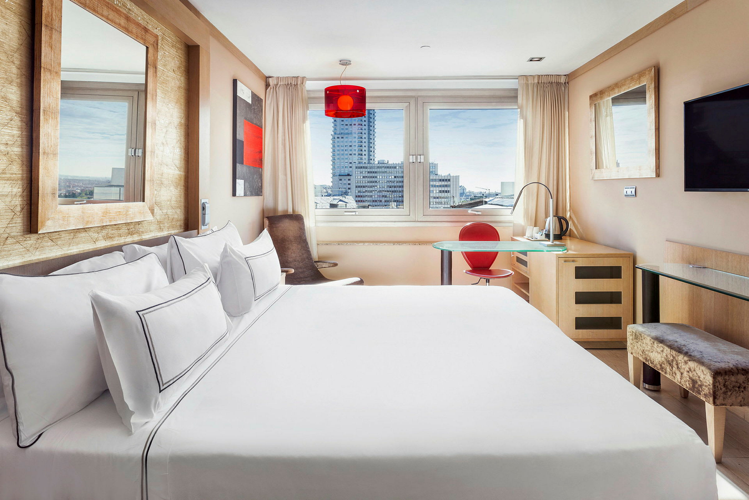 Valoisa ja moderni hotellihuone, jossa on suuri sänky, ikkuna, josta näkyy kaupungin rakennuksia, työpöytä punaisella tuolilla ja seinälle kiinnitetty televisio. (Tekoälyn luoma teksti)