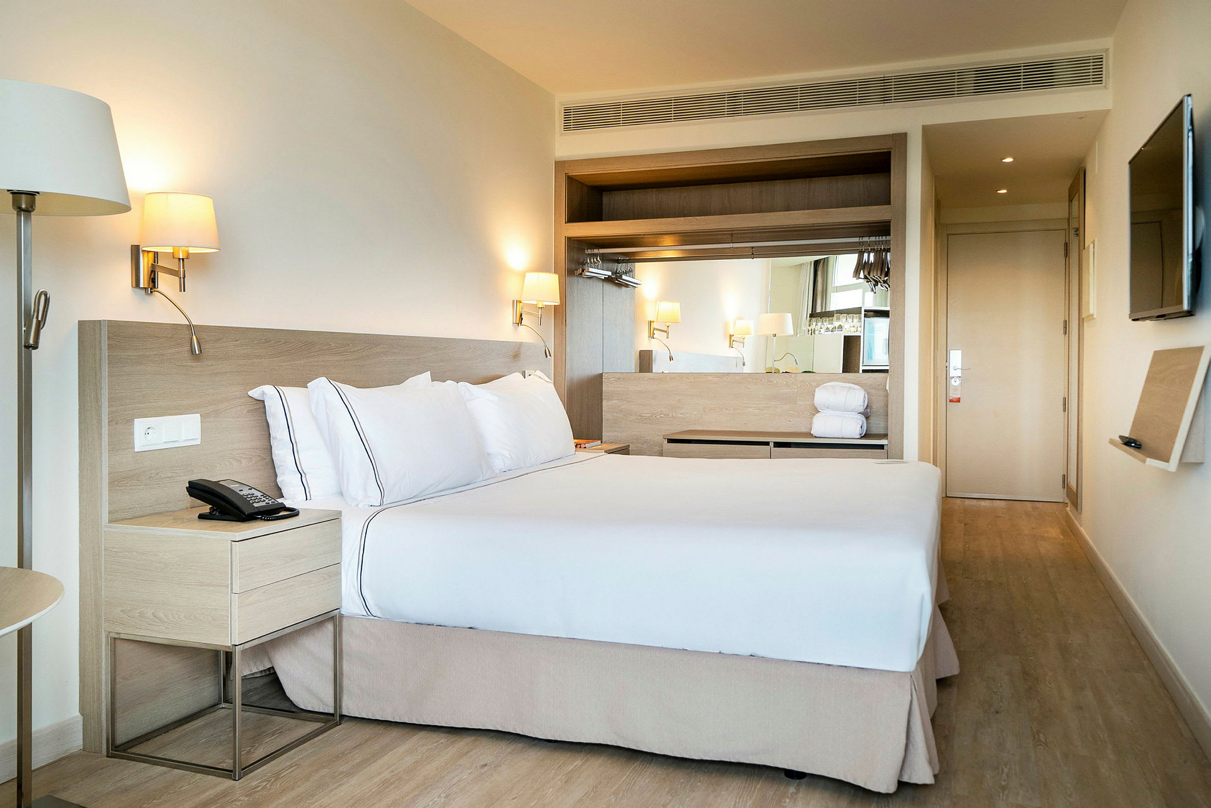 Hotellihuone, jossa on suuri sänky ja valkoiset liinavaatteet sekä tyynyt. Lampuja roikkuu sängyn sivuilla ja yöpöytä on sängyn vieressä. (Tekoälyn luoma teksti)