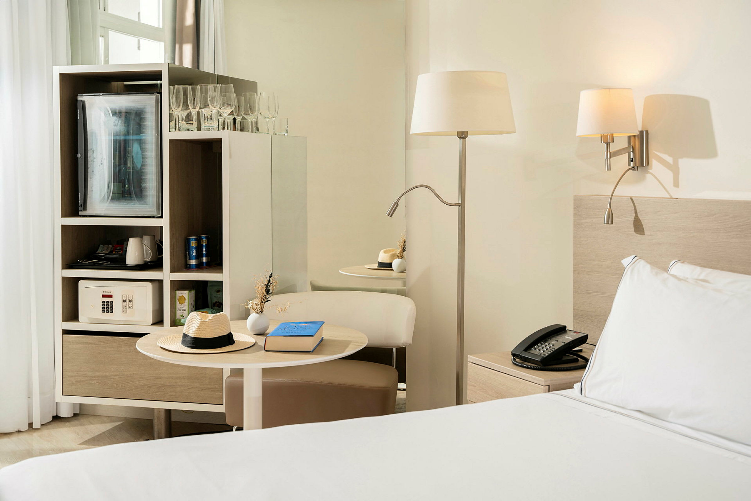 Hotellihuone, jossa on sänky yöpöydän vieressä, pöytälamppu ja sisäänrakennettu huonekalu, johon kuuluu jääkaappi ja kassakaappi. (Tekoälyn luoma teksti)