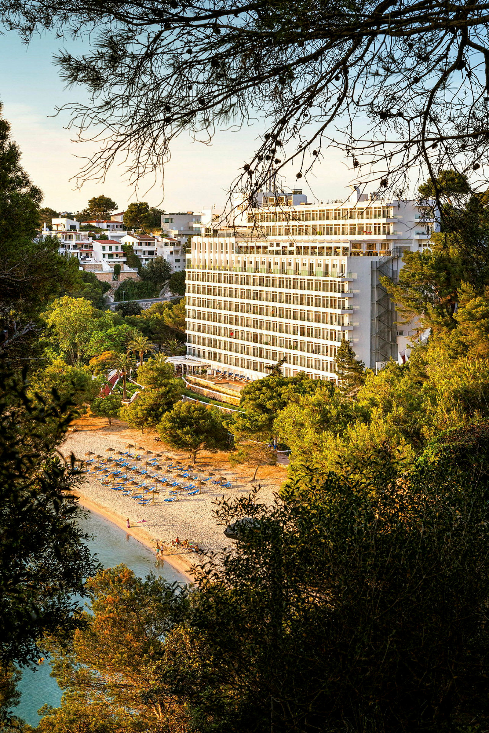 Kuva hotellista, joka sijaitsee lähellä rantaa aurinkotuoleilla ja aurinkovarjoilla, ympäröitynä vehreillä puilla. (Tekoälyn luoma teksti)