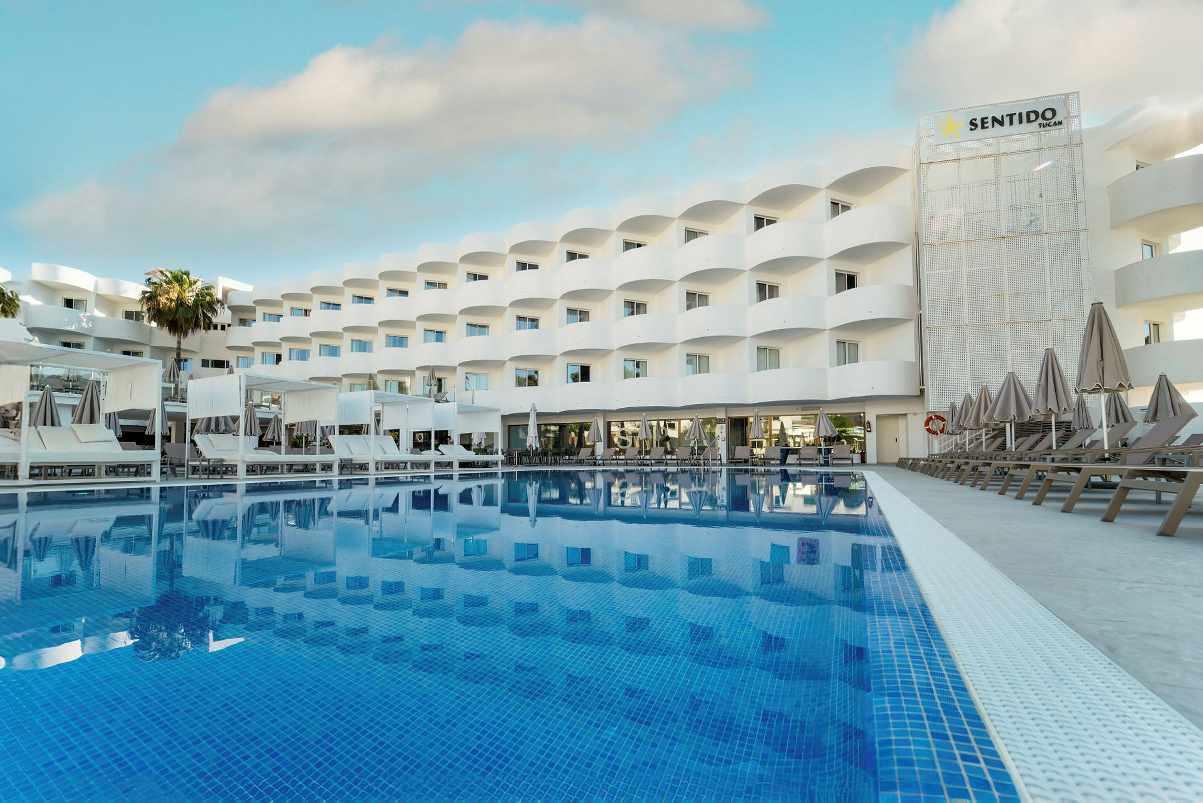 Kuva hotellista, jonka edessä on suuri uima-allas. Rakennus on valkoinen ja siinä on parvekkeita sekä aurinkotuoleja altaan äärellä. (Tekoälyn luoma teksti)