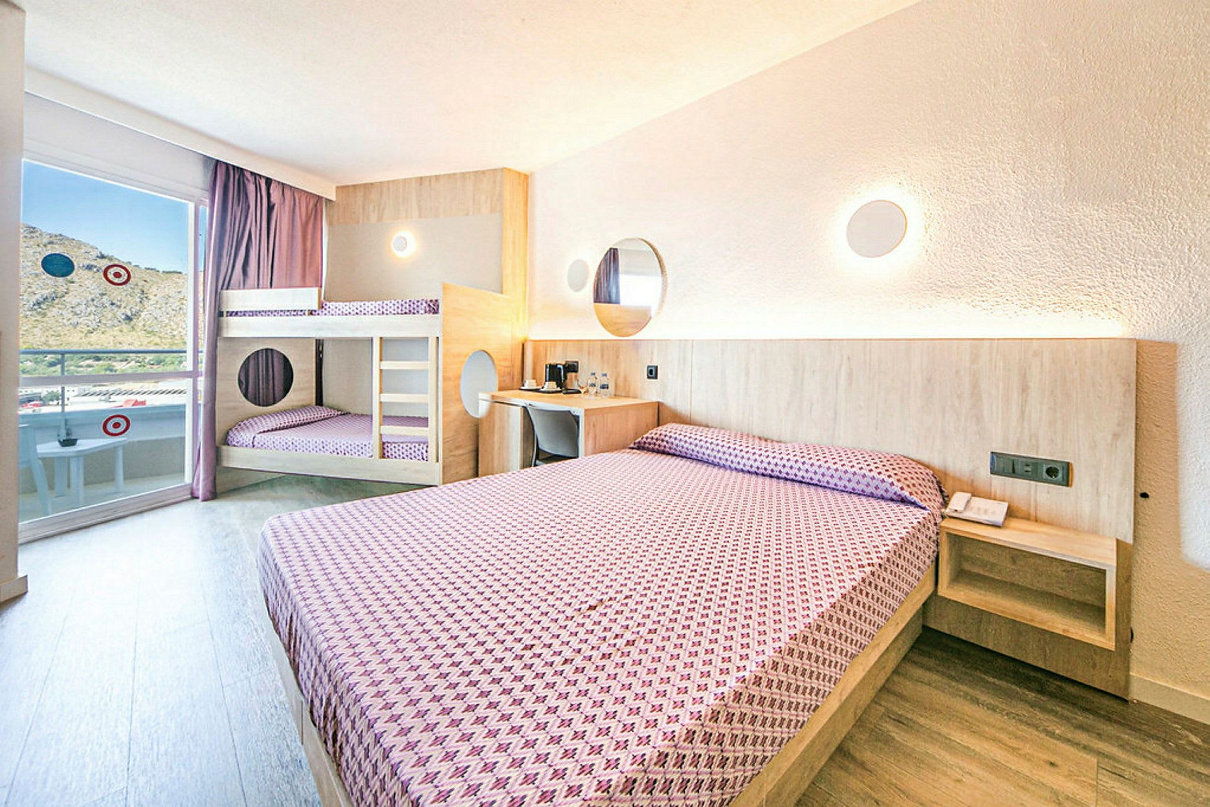 Hotellihuone, jossa on parivuode ja kerrossänky, molemmat pedattu violetilla kuvioidulla vuodevaatteella, vaaleat seinät ja puuyksityiskohtia. (Tekoälyn luoma teksti)