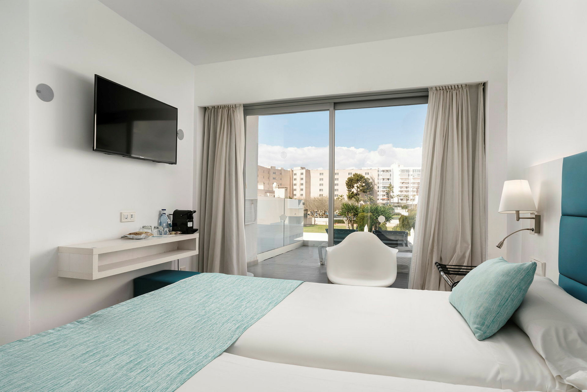 Hotellihuone, jossa kaksi vuodetta vaaleilla lakanoilla ja turkooseilla päiväpeitteillä. Tuoli on lasioven edessä, joka johtaa parvekkeelle. Seinään kiinnitetty televisio on hyllyn vieressä, jossa on vesipulloja ja kahvinkeitin. (Tekoälyn luoma teksti)