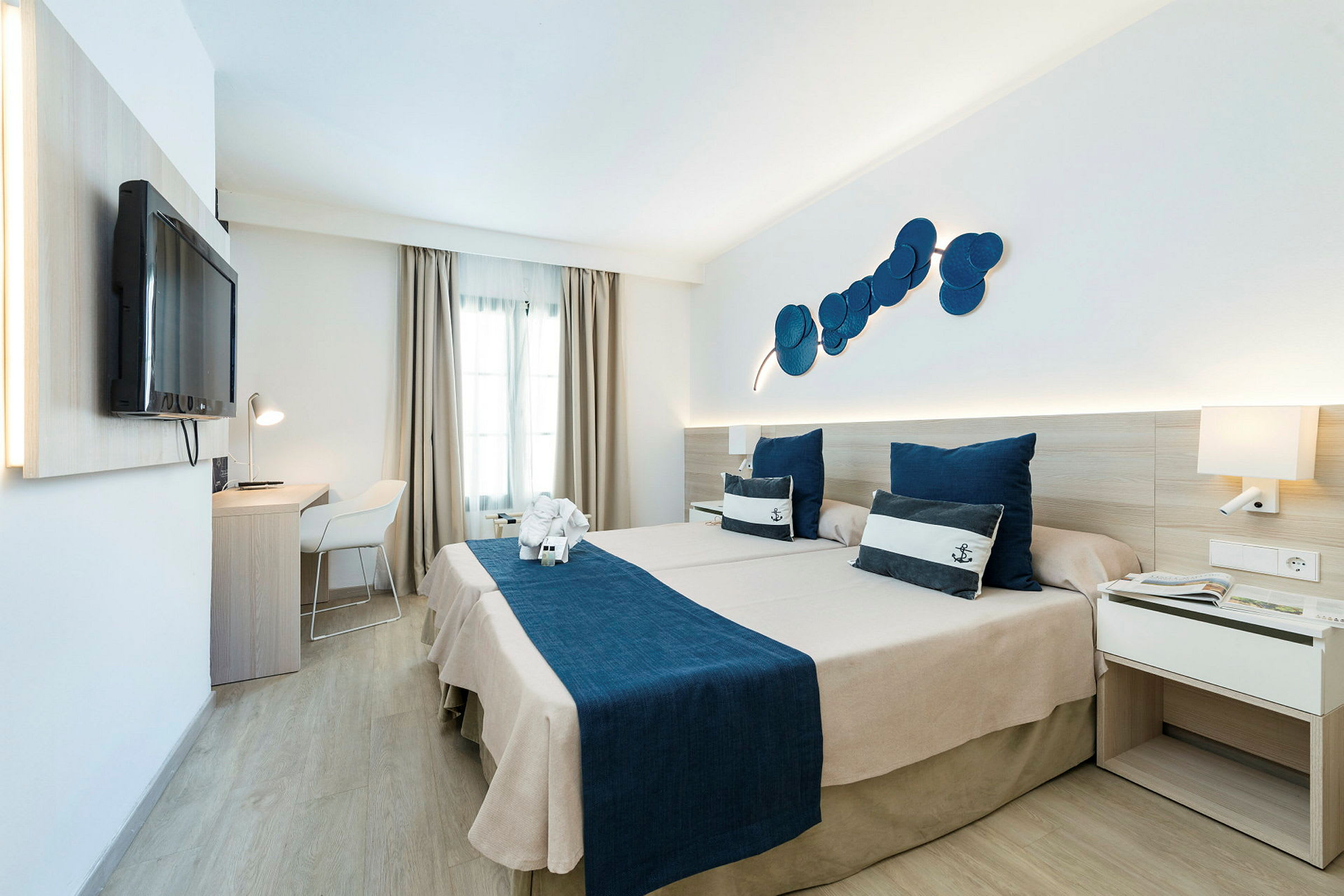 Hotellihuone, jossa kaksi sänkyä ja merellinen teema, siniset vuodevaatteet ja koristeelliset tyynyt. (Tekoälyn luoma teksti)