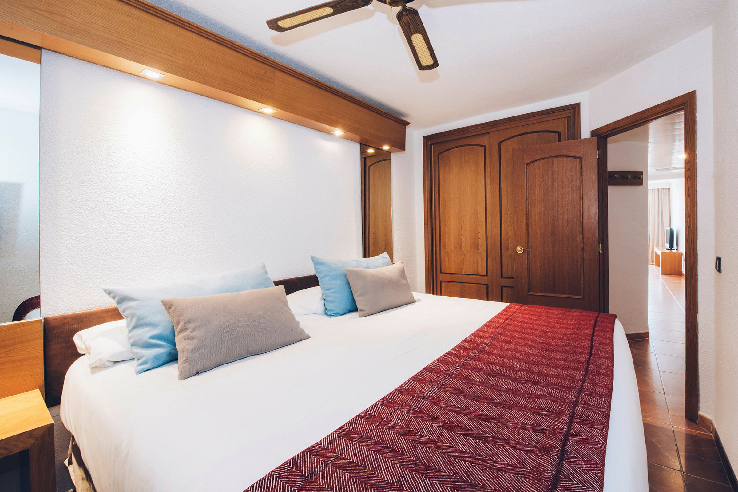 Hotellihuone, jossa on iso sänky, tyynyjä ja punainen päiväpeite. Huoneessa on kattotuuletin ja puuovilla varustettu kaappi. (Tekoälyn luoma teksti)