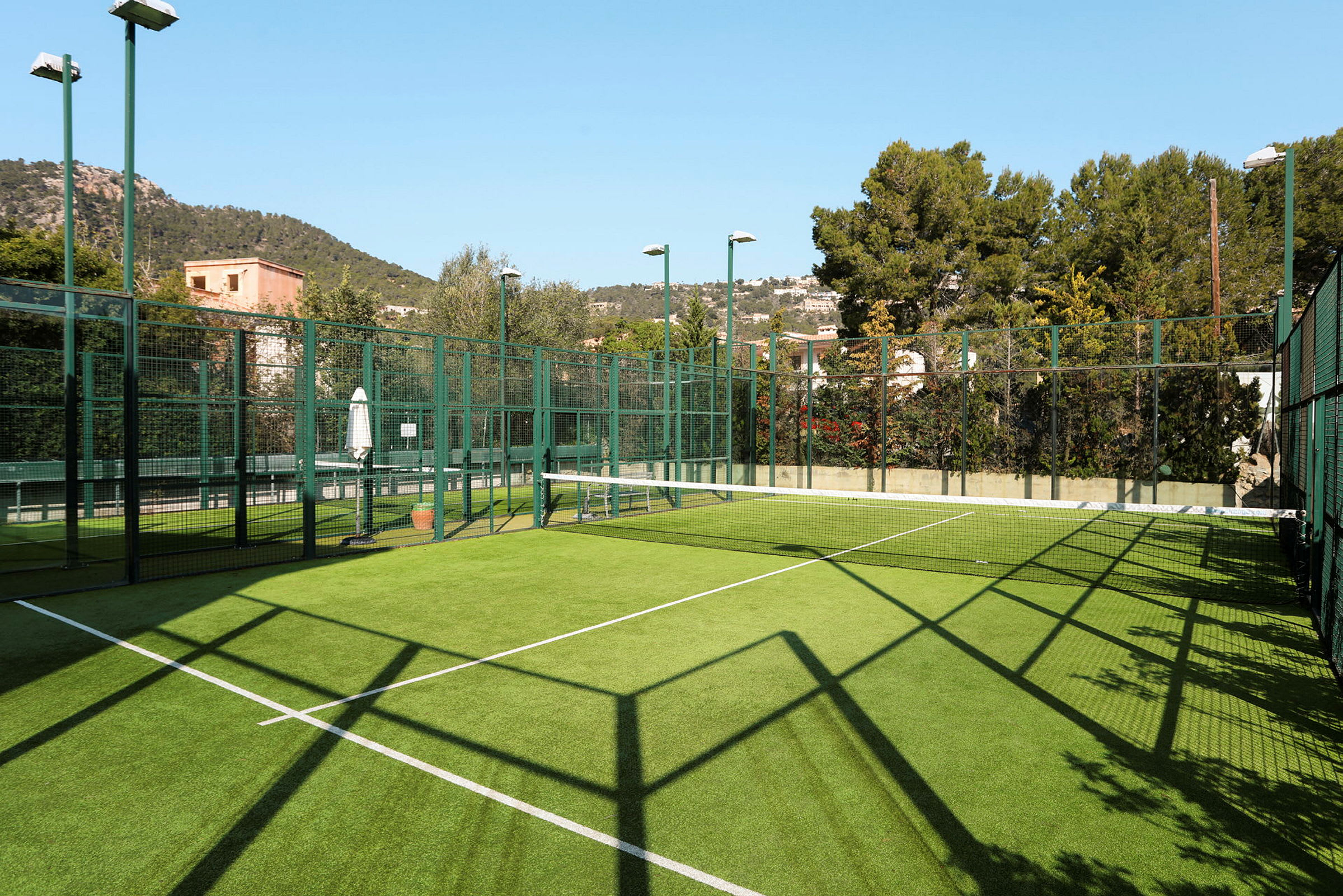 Padel-kenttä ulkona, jossa on vihreä keinonurmi, ympärillä turvaverkkoja sekä puita ja rakennuksia. (Tekoälyn luoma teksti)