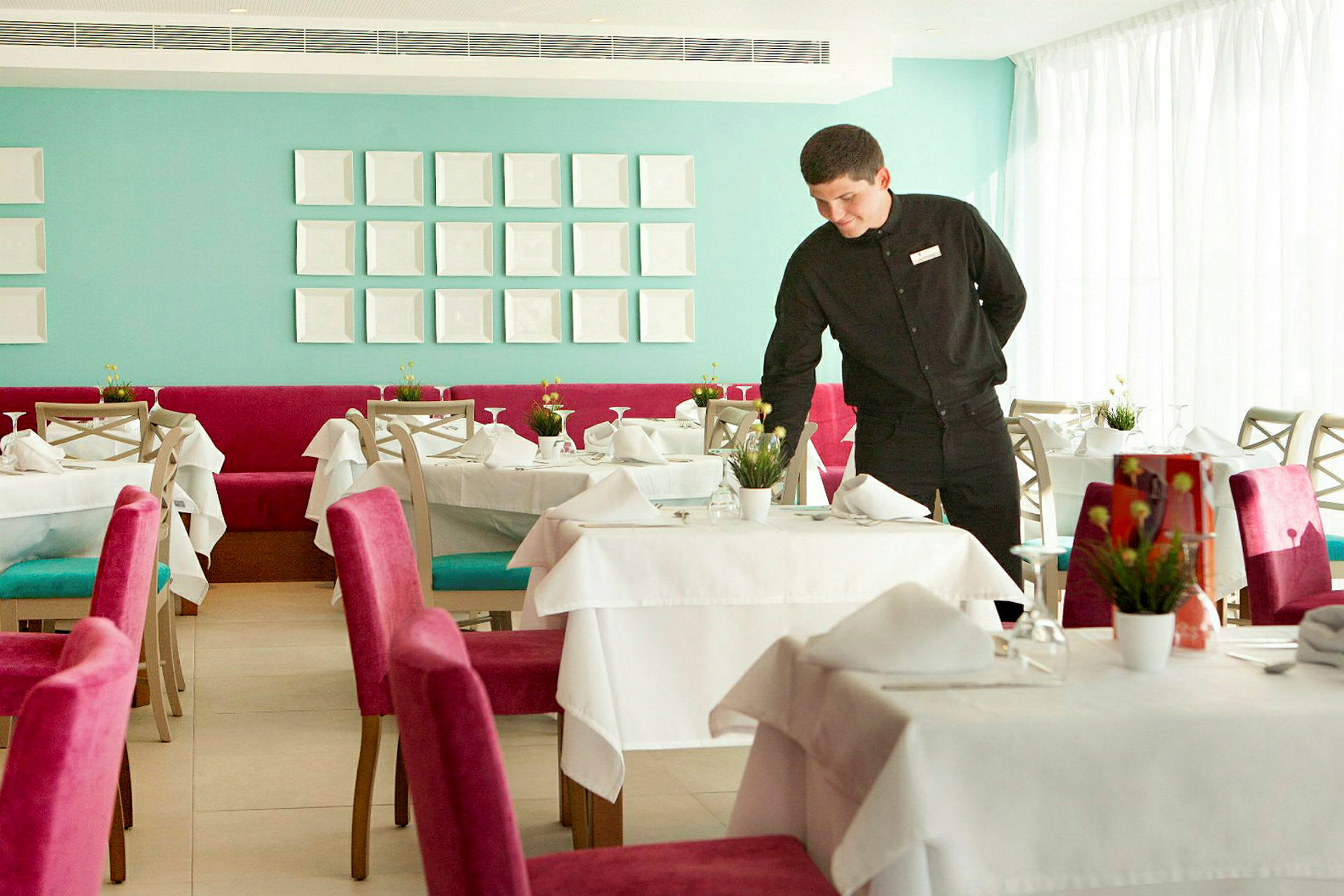 Tyylikäs ravintola, jossa on katetut pöydät ja värikkäitä tuoleja, tarjoilija valmistautuu pöytään. (Tekoälyn luoma teksti)