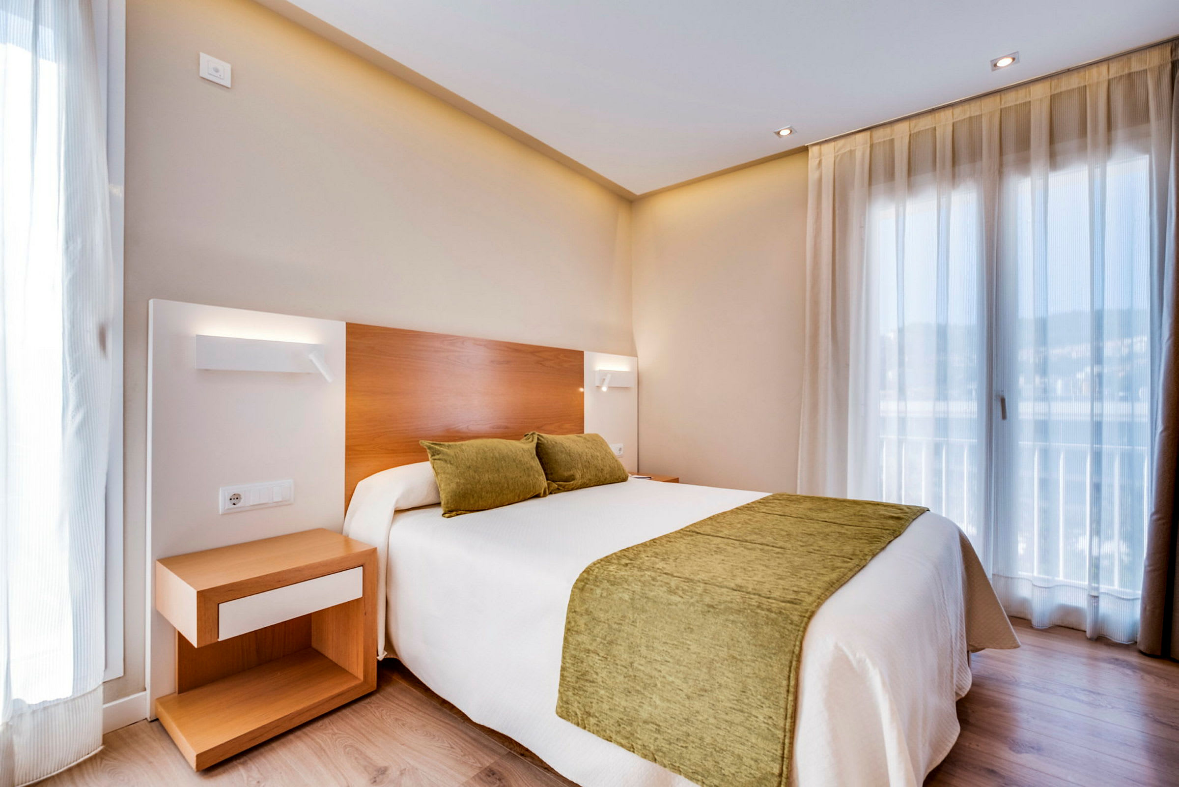 Hotellihuone, jossa on kaksi vihreää tyynyä ja peitto valkoisella sängyllä. Yöpöytä sijaitsee sängyn vieressä. (Tekoälyn luoma teksti)