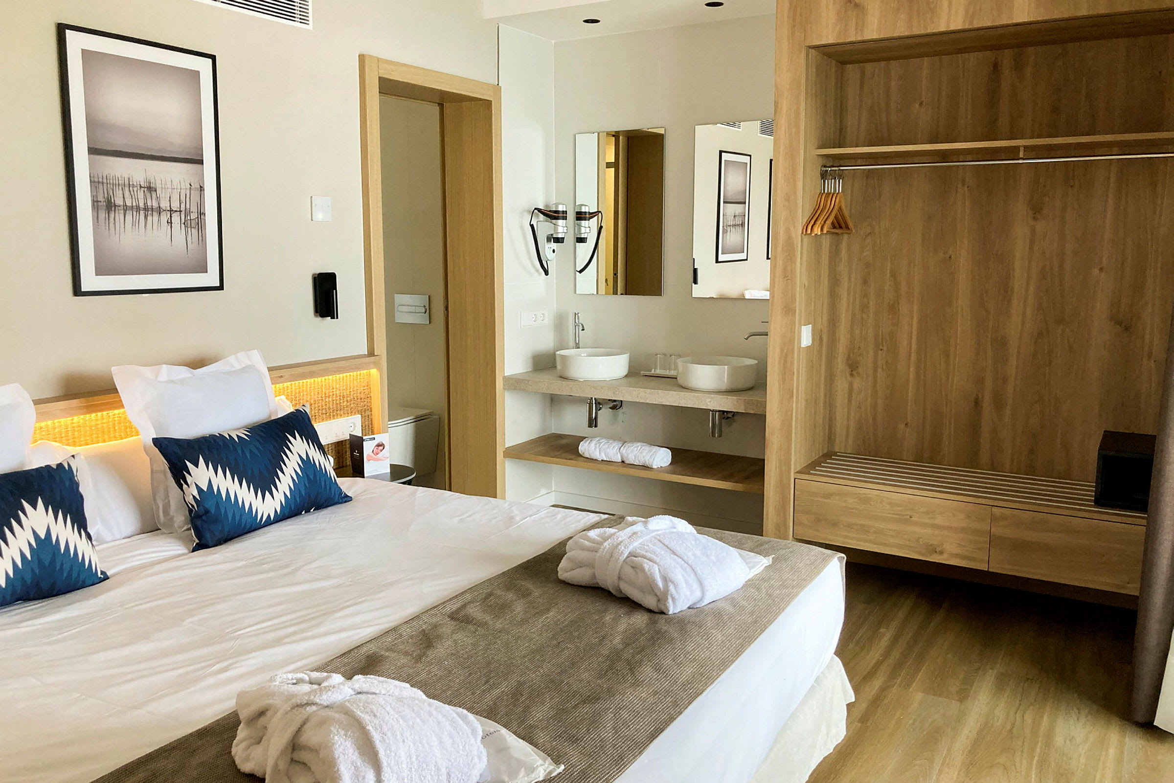 Hotellihuone, jossa on parivuode, kaksi sinistä tyynyä ja valkoiset kylpytakit sängyllä. Taustalla on kylpyhuone, jossa on kaksi pesuallasta ja peili sekä puupintainen vaatekaappi. (Tekoälyn luoma teksti)