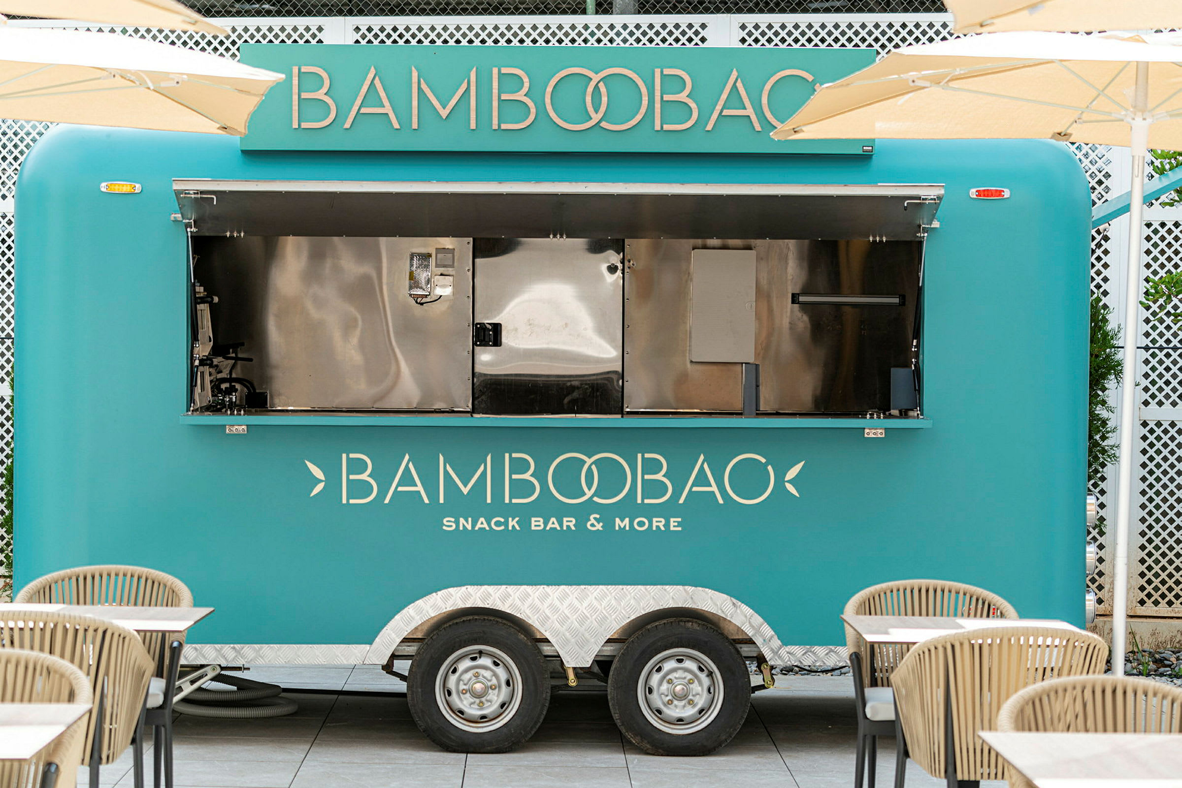 Turkoosinvärinen ruokavaunu, jossa lukee 'Bamboobao Snack Bar & More', on pysäköity pöytien ja tuolien ympäröimänä. (Tekoälyn luoma teksti)