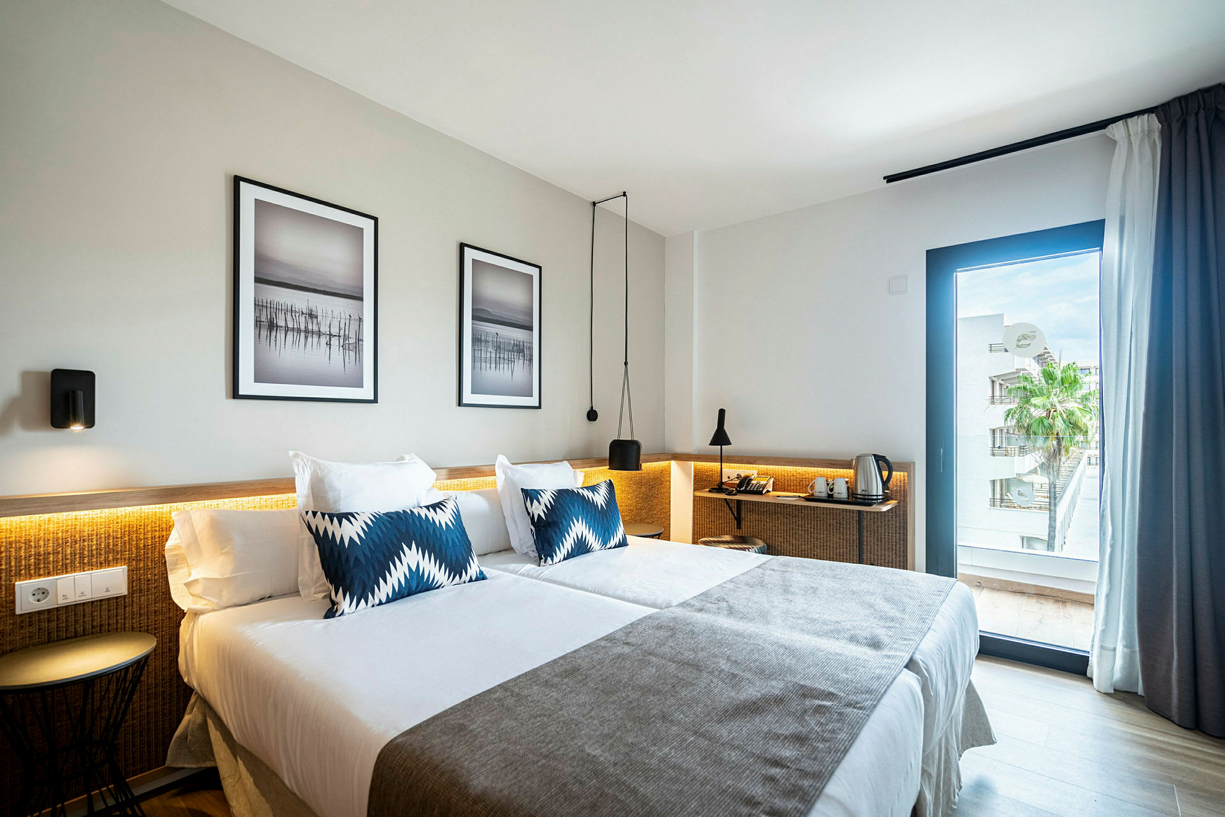 Hotellihuone, jossa on kaksi sänkyä, peitetty valkoisilla vuodevaatteilla ja harmailla peitteillä. Sängyillä on siniset koristetyynyt. Seinillä on kaksi kehystettyä taideteosta, ja ikkuna tarjoaa näkymän palmupuun ja rakennusten yli. (Tekoälyn luoma teksti)