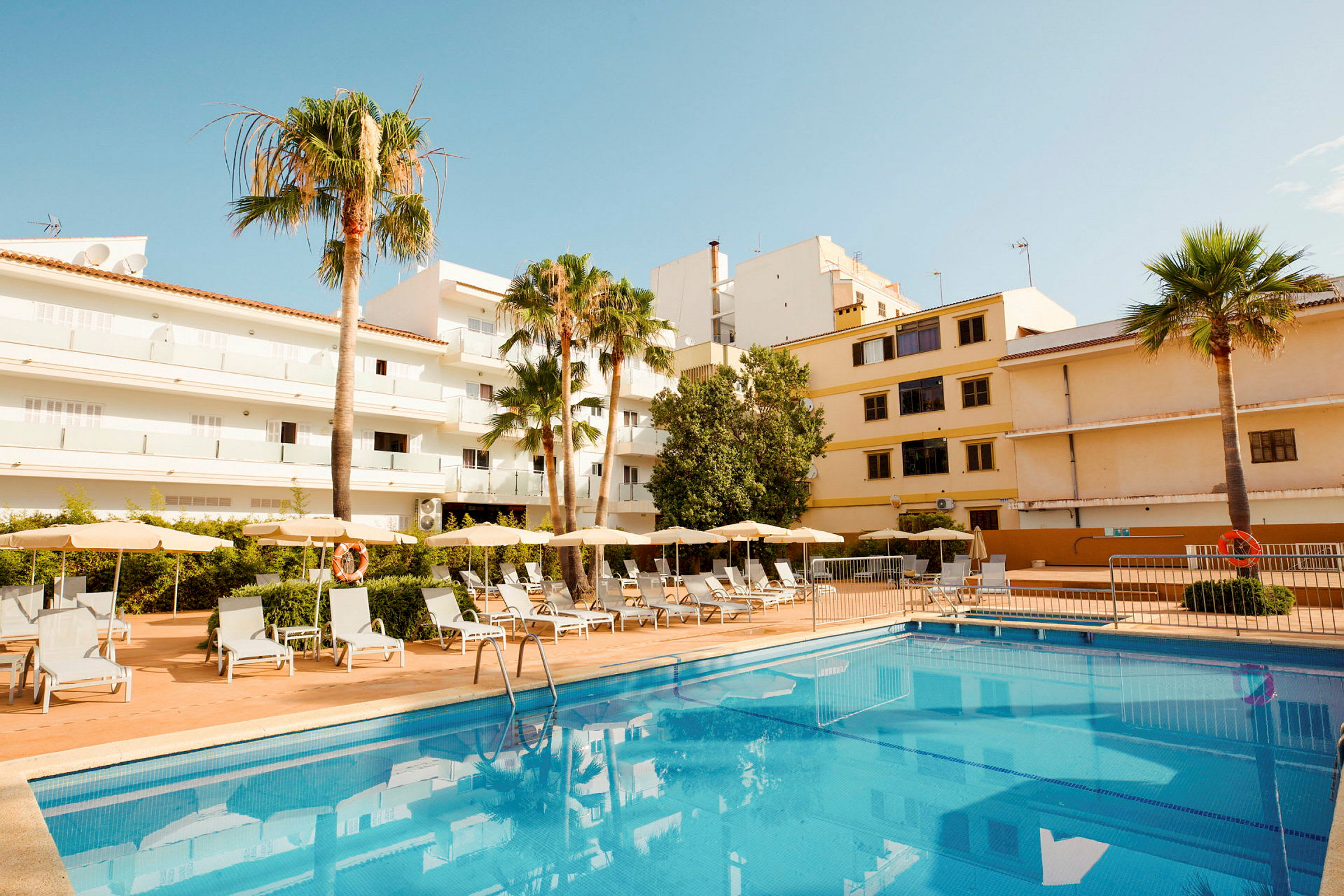 Hotelli ulkopuolelta, jossa uima-allas ja aurinkotuoleja, rakennusten ja palmujen ympäröimänä. (Tekoälyn luoma teksti)