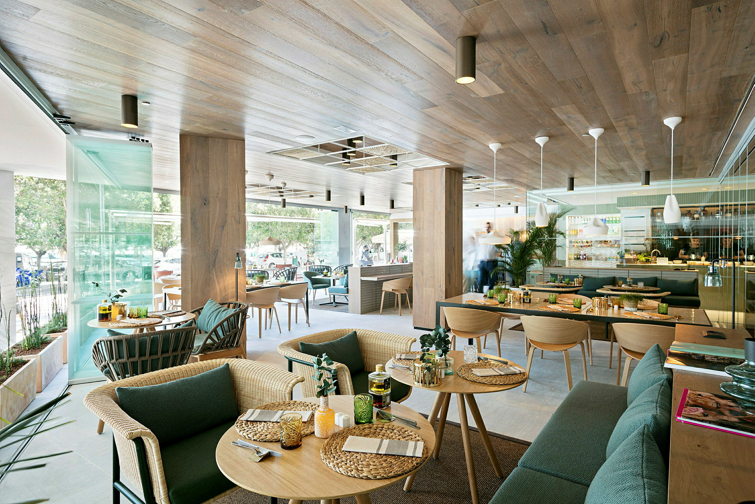 Moderni ravintola, jossa on puusisustus ja suuret lasi-ikkunat. Pöydillä on tarjoiltavaa ruokaa ja sisustuksessa on käytetty vihreitä kasveja. (Tekoälyn luoma teksti)