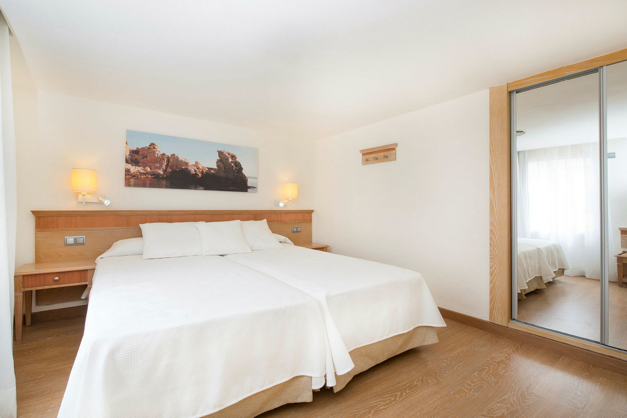 Hotellihuone, jossa on kaksi erillistä vuodetta, valkoiset vuodevaatteet, vuoteiden takana puupaneeli pöytälampuilla, kaapin peiliovet ja suuri ikkuna, josta tulvii luonnonvaloa. (Tekoälyn luoma teksti)